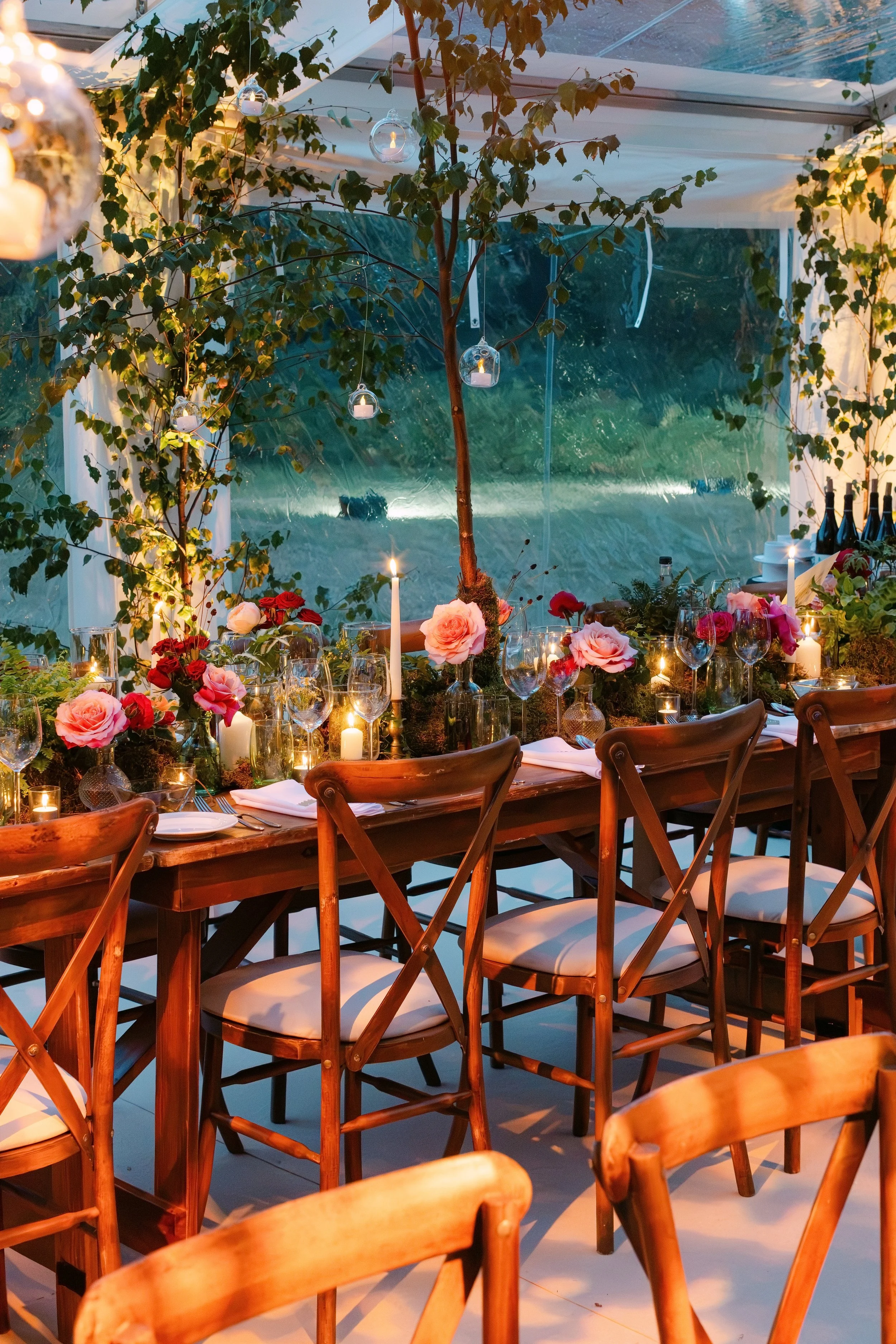 A beautifully decorated dinner table inside a tent with string lights, candles, and flowers, with a view of greenery outside.