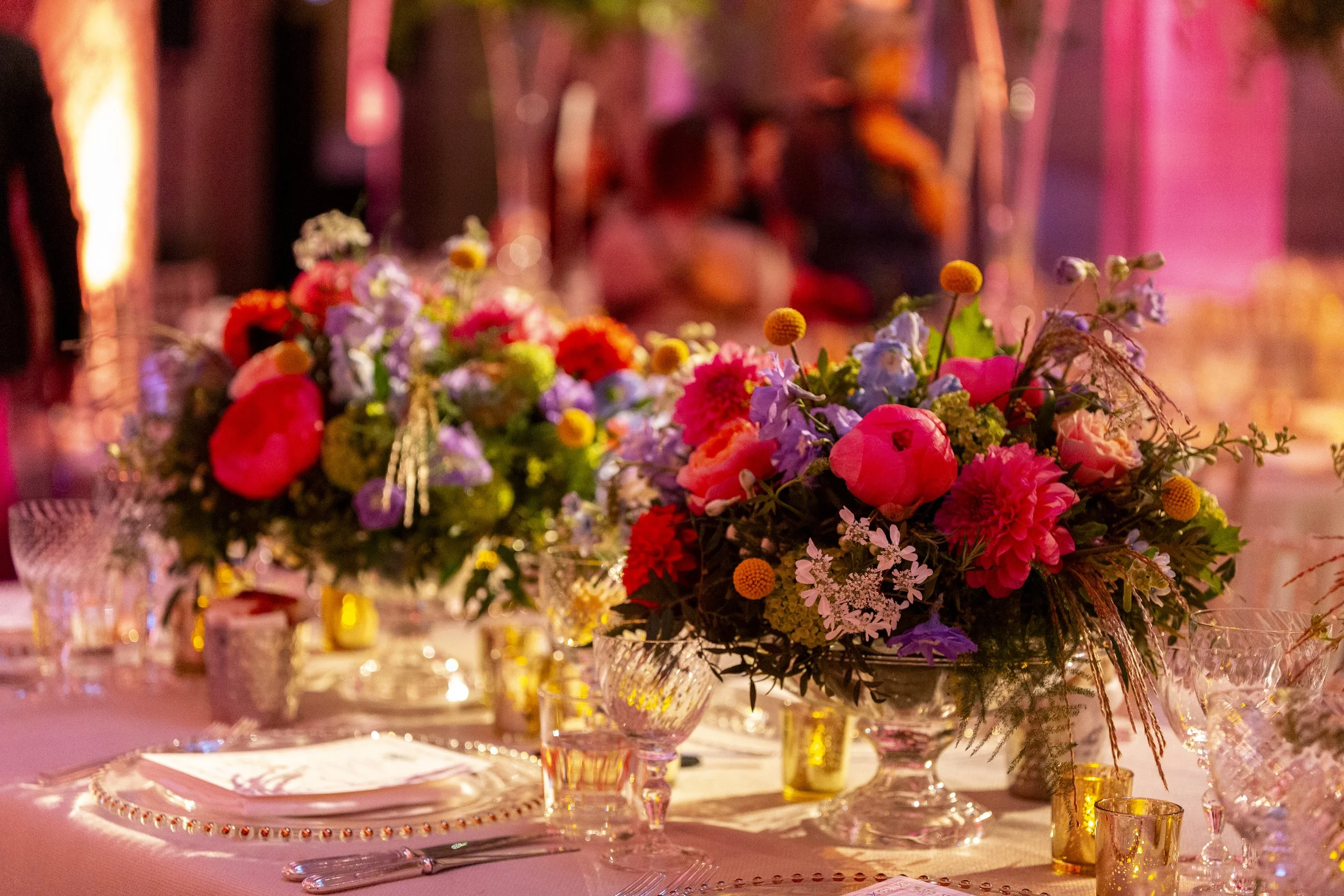 A beautifully decorated table with a large floral arrangement as a centerpiece, surrounded by glassware, plates, and gold-colored votive candles, set for an event in a warmly lit venue.