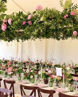 Wedding reception table decorated with pink and white flowers, greenery, and candles, under a floral canopy.