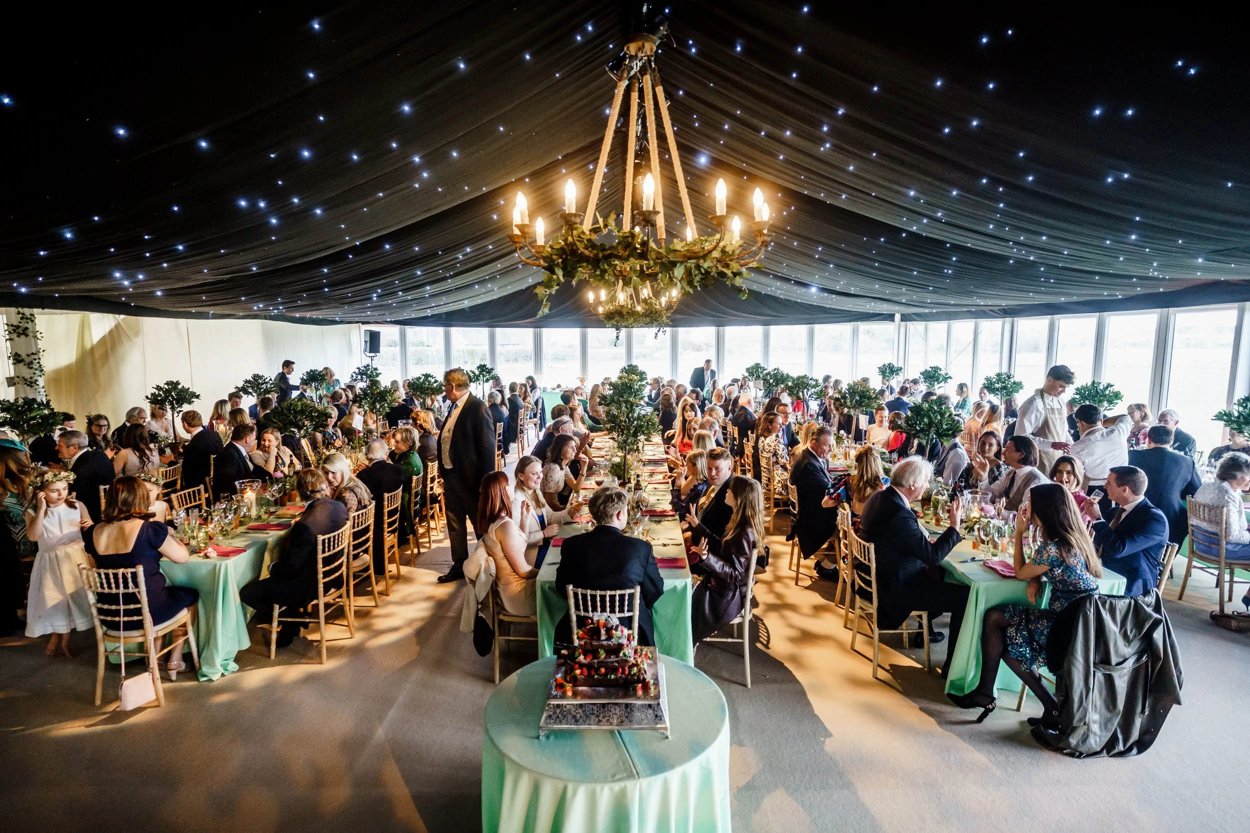 A large banquet hall filled with people seated at long tables decorated with greenery and flowers for a celebration. There is a majestic chandelier hanging from a dark, starry ceiling. The hall has large windows that let in natural light, and a buffe