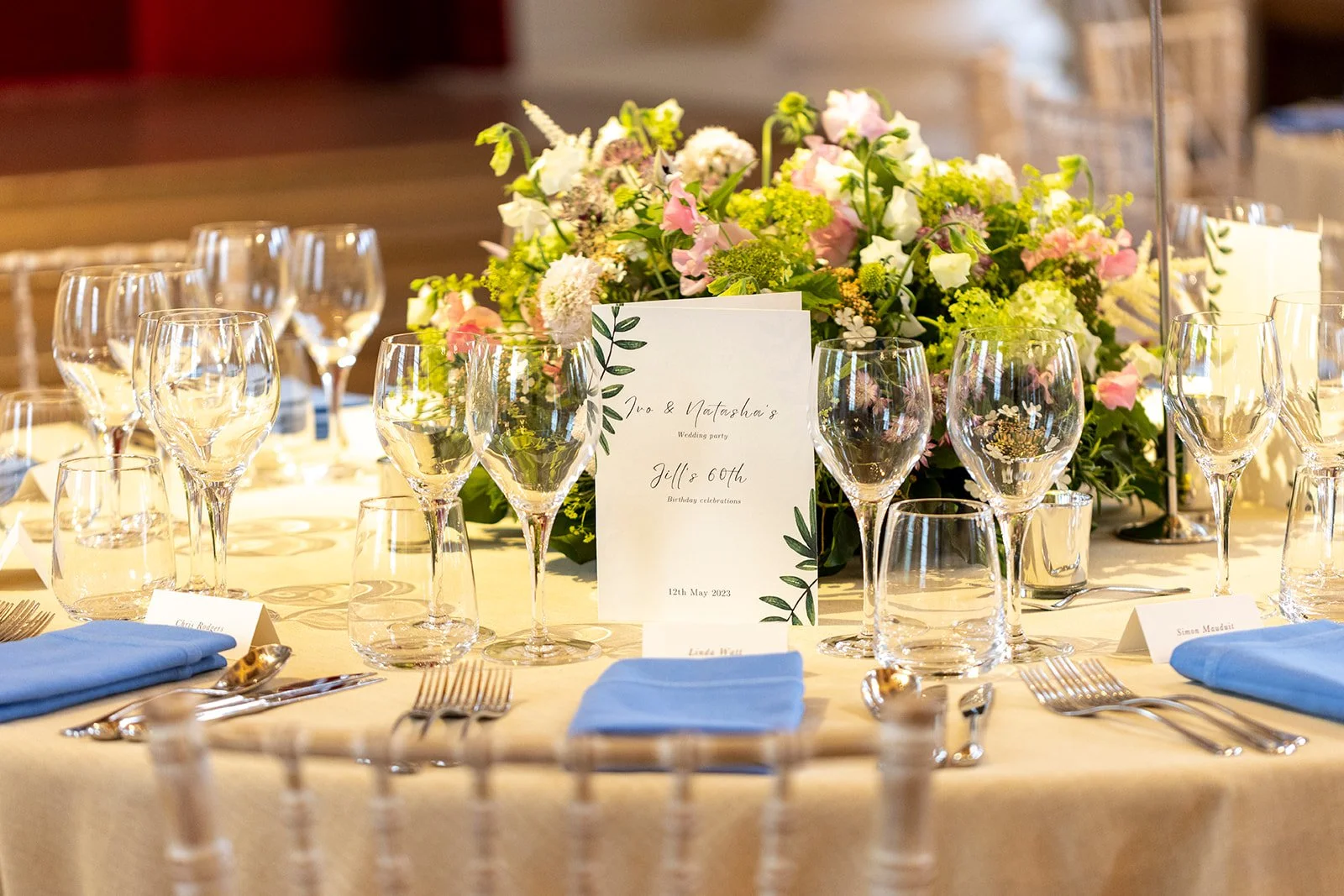 Wedding reception table with elegant floral centerpiece, multiple glasses of water and wine, silverware, light blue napkins, and a wedding program card reading 'Iva & Natasha's Wedding party, Jill's 60th Birthday celebrations, 12th May 2023'.