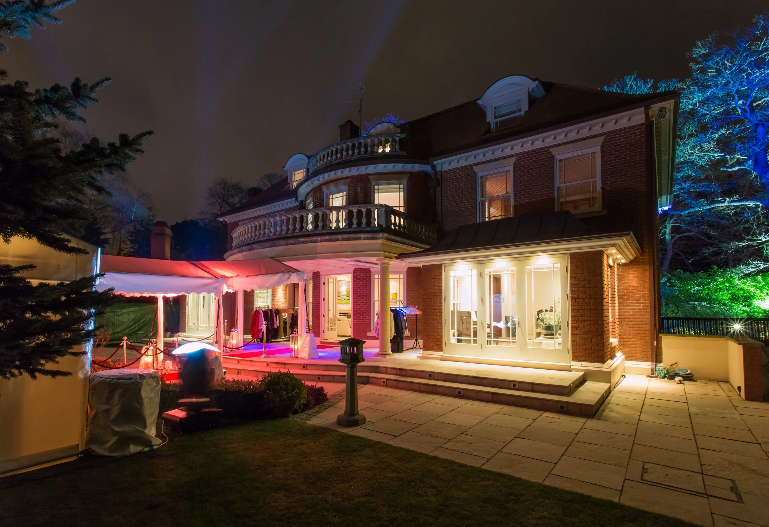 Night view of a large brick house with illuminated windows, a porch, and a balcony, decorated with colorful lighting. There is an outdoor tent, clothing rack, and garden area with outdoor lights.