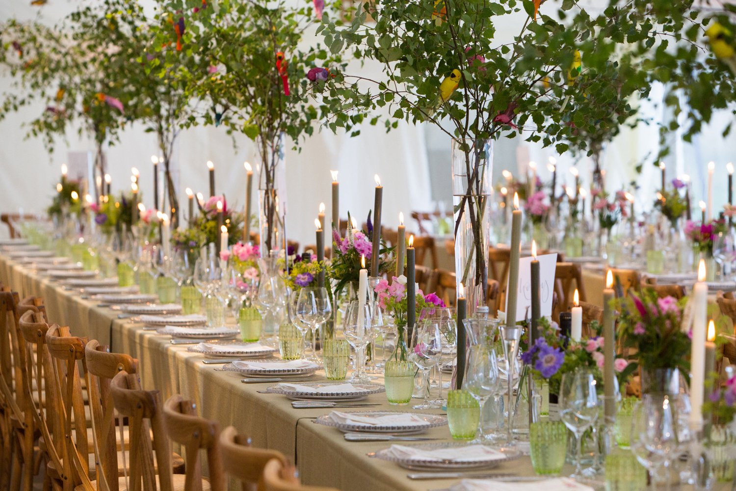 A long banquet table decorated with pink, purple, and green flowers, tall candles, and glassware, with trees and candles above in a bright indoor setting.