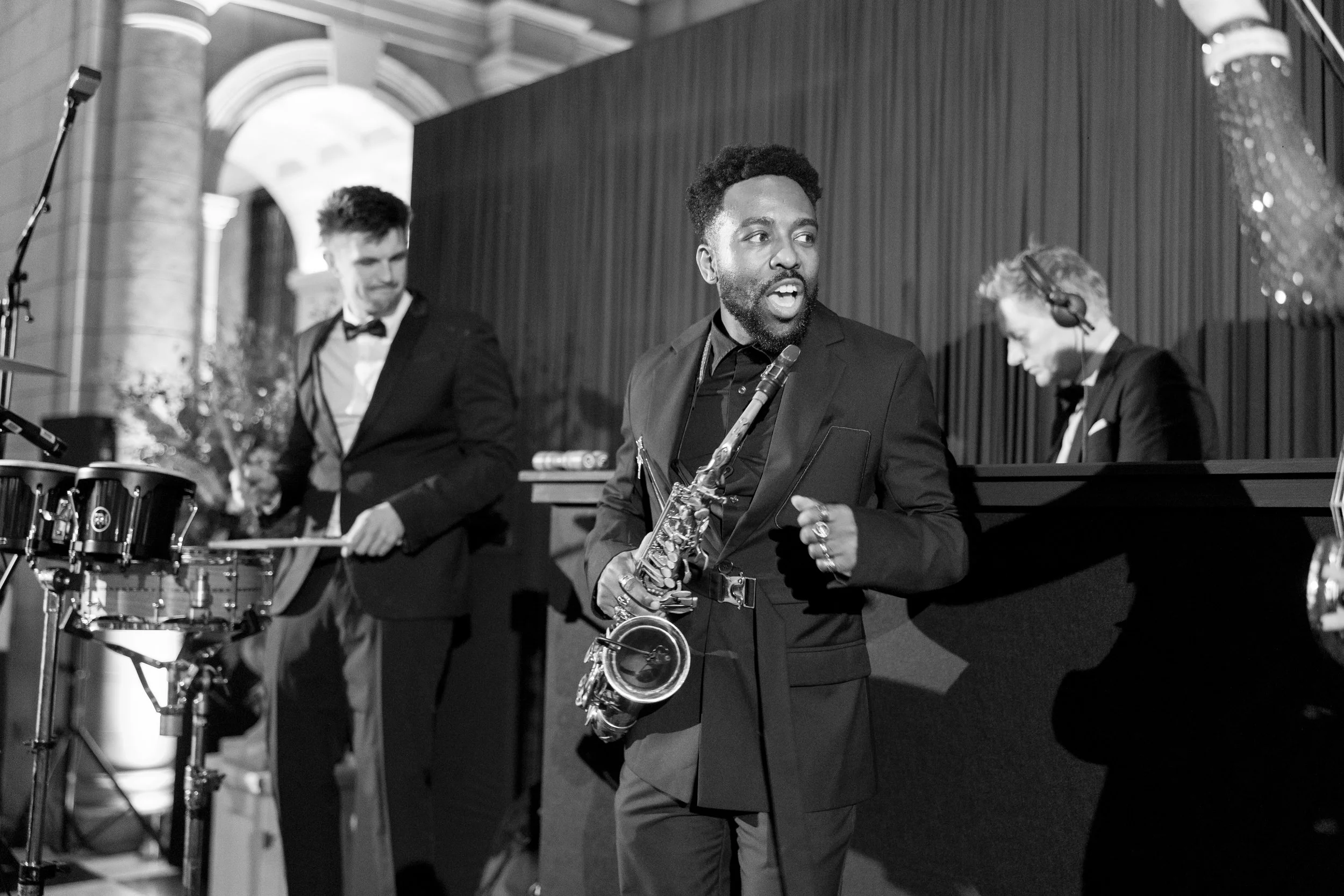 A group of three male musicians performing on stage, with one playing drums, one singing into a microphone, and another at a keyboard. The setting appears to be an indoor venue with decorative arches in the background.