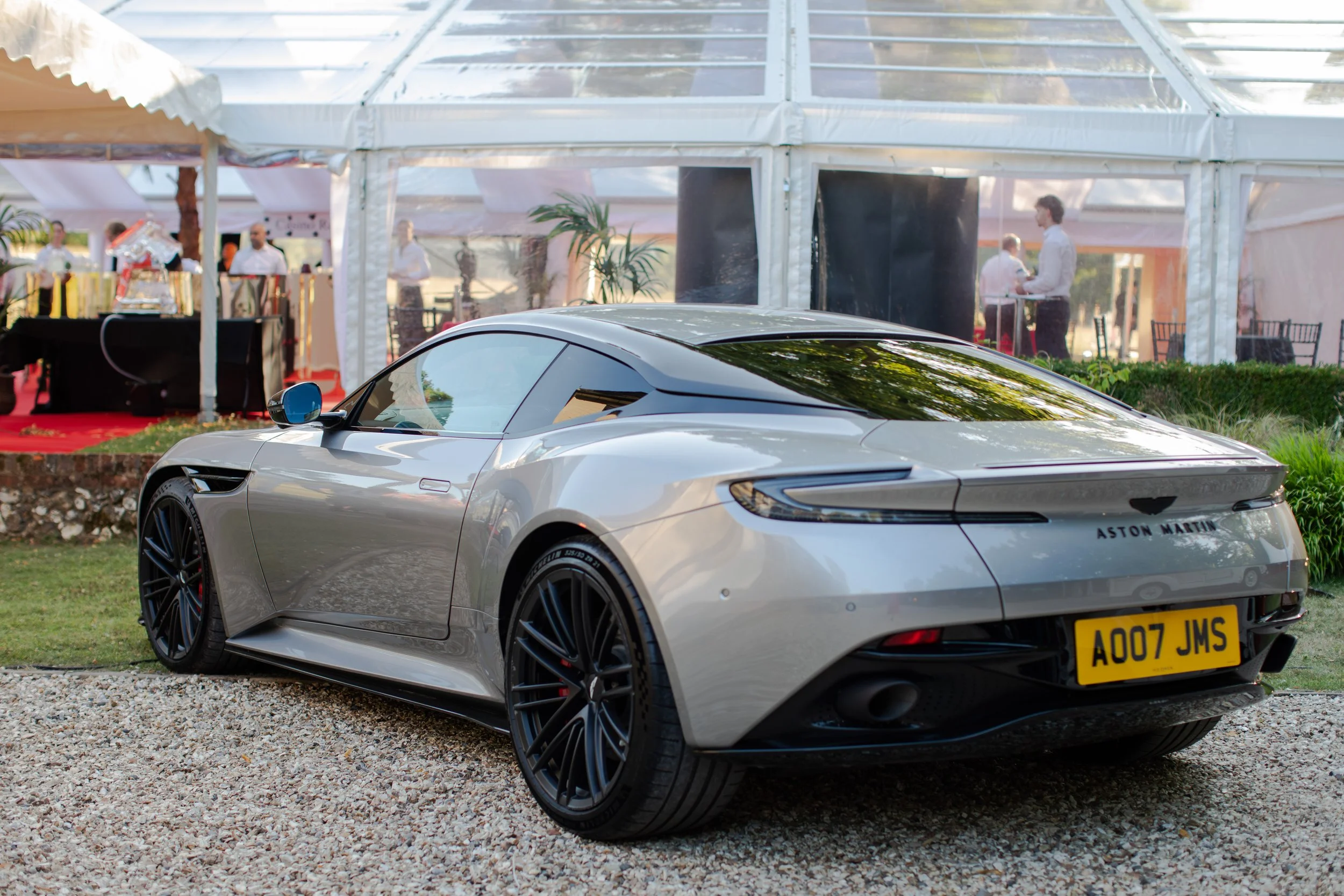 A silver Aston Martin sports car parked on a gravel surface at an outdoor event. In the background, there is a white tent with people inside, some standing and talking. The scene is during daylight.