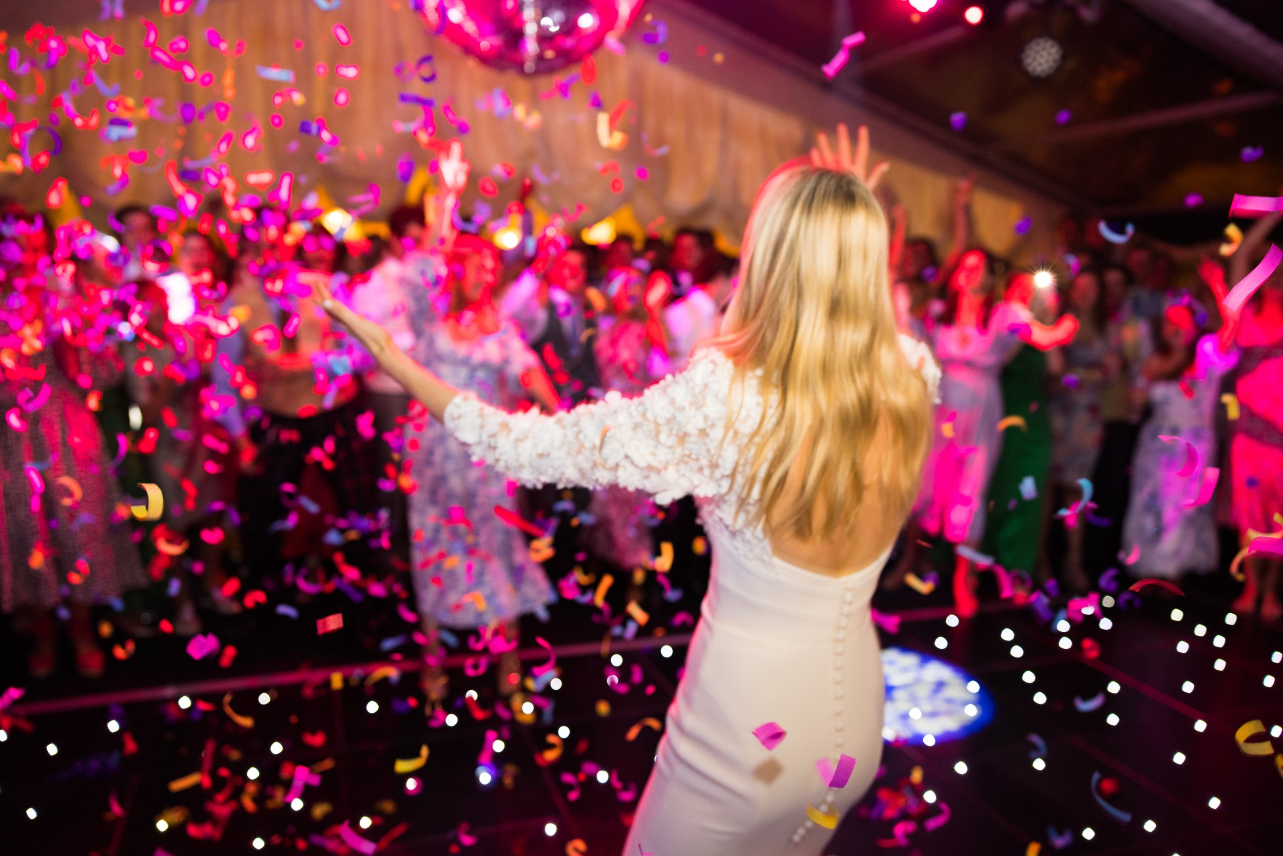 A woman with long blonde hair wearing a white dress at a celebration or party, surrounded by colorful confetti falling in the air, with a crowd of people in the background.