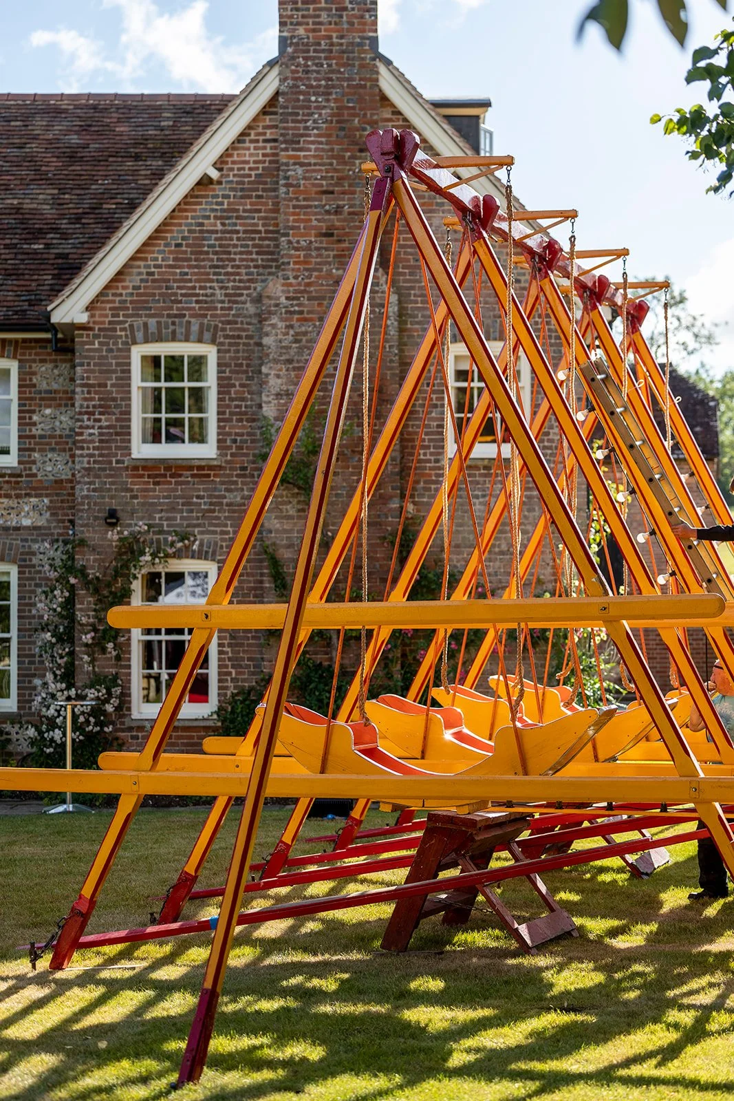 A yellow and red wooden swing set with multiple swings in a backyard with a brick house in the background.