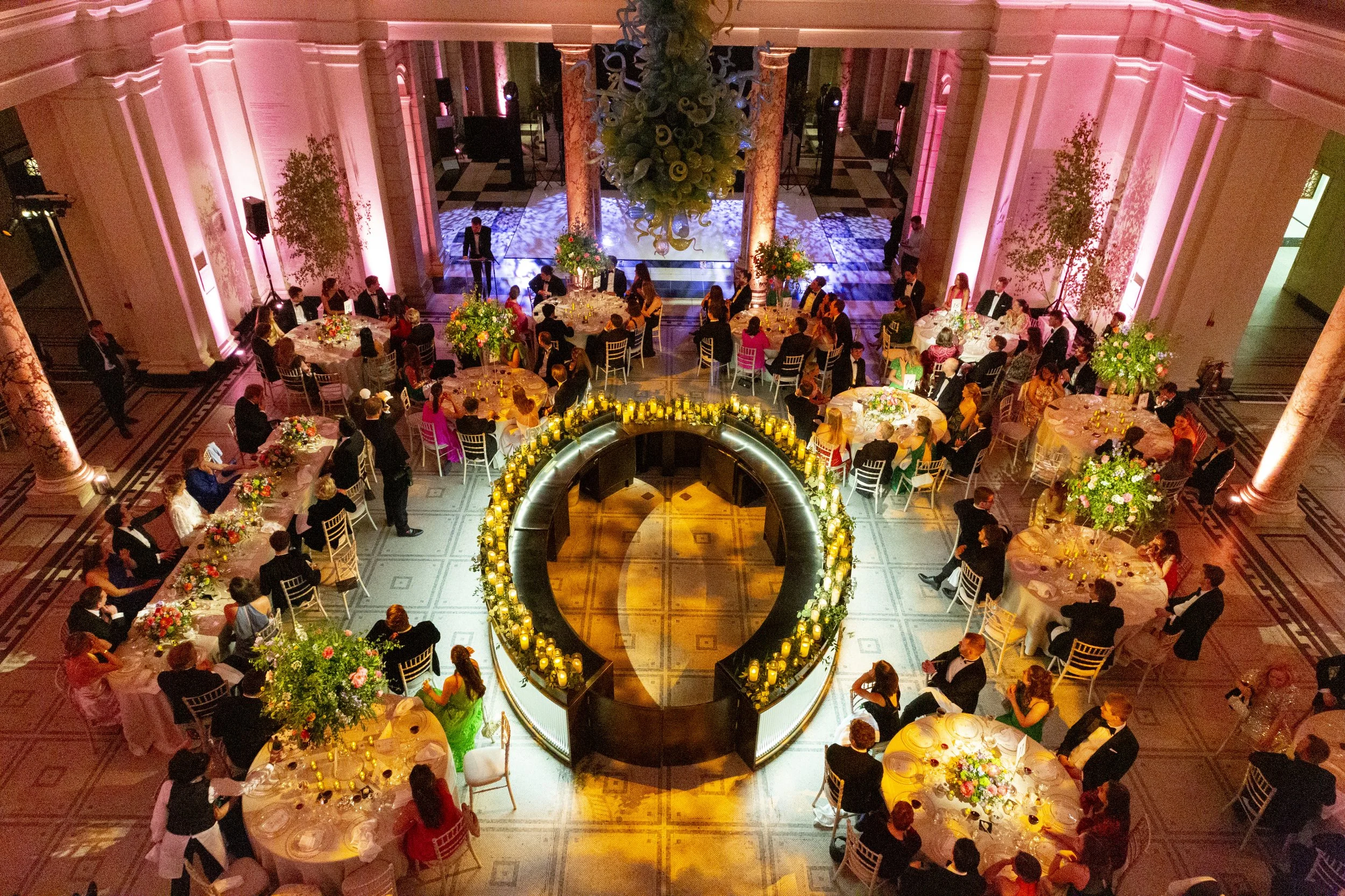 A large banquet hall filled with round tables decorated with floral centerpieces, surrounded by guests wearing formal attire, with a circular bar at the center adorned with candles and greenery, and a stage area with lighting in the background, pink uplighting on the walls, and a large hanging floral arrangement overhead.