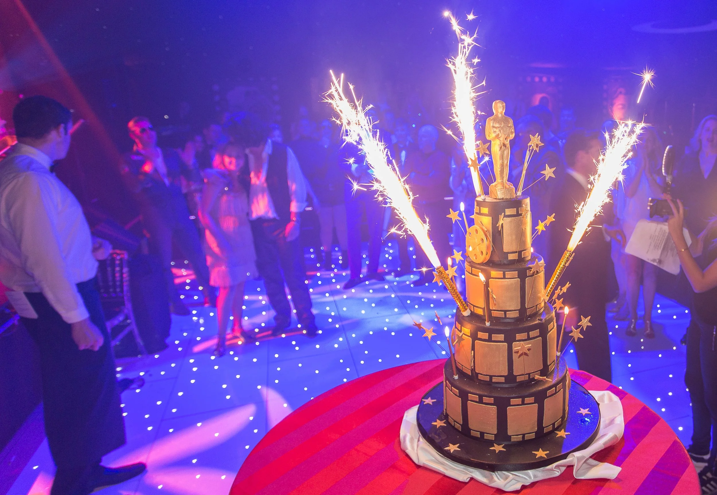 A large multi-tiered cake decorated with a castle design, surrounded by sparklers, with party guests in formal attire in the background, celebrating at a party or event.