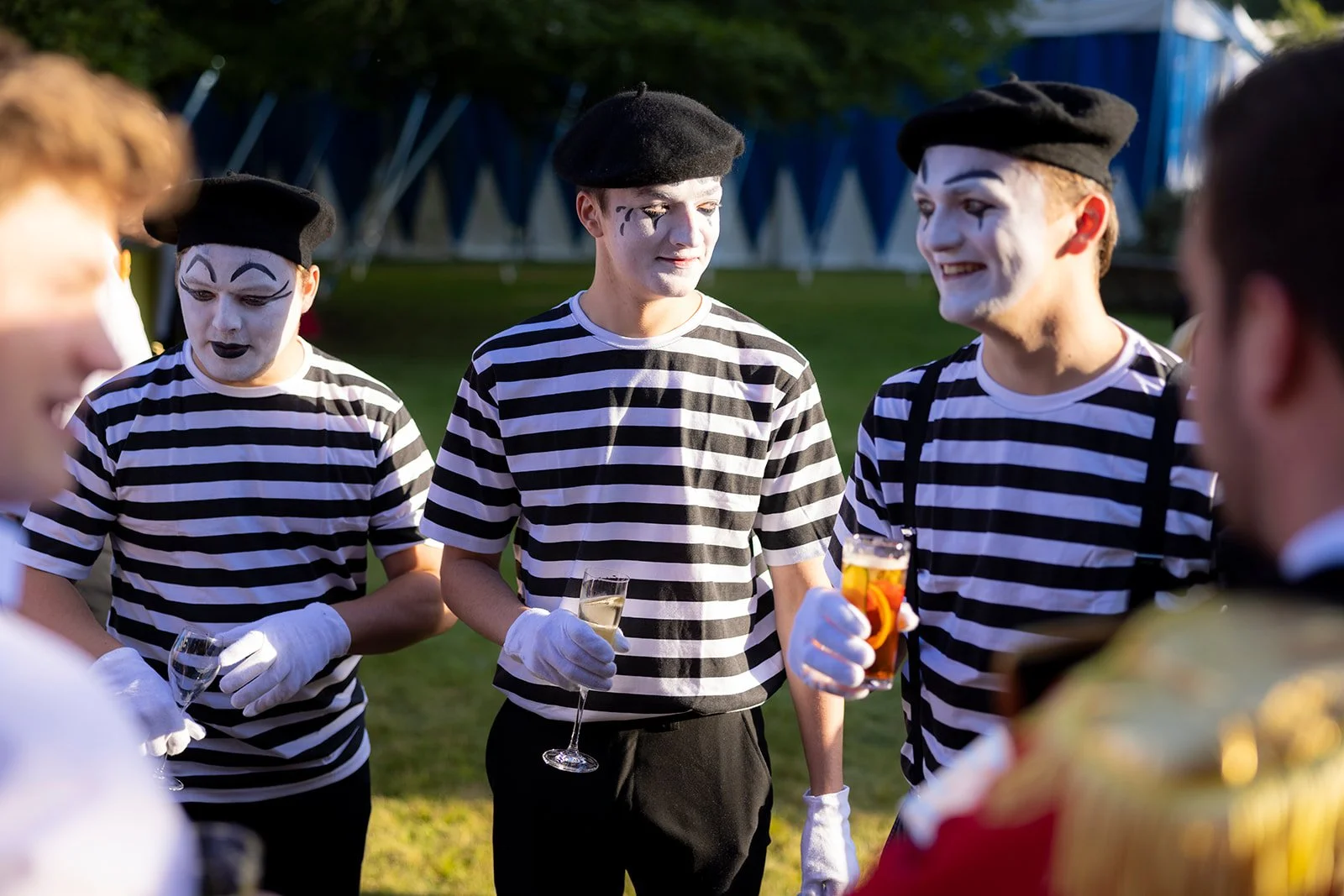 Group of young people dressed as mime artists in striped shirts, black berets, and white gloves, standing outdoors at a gathering or event, holding drinks and engaging in conversation.