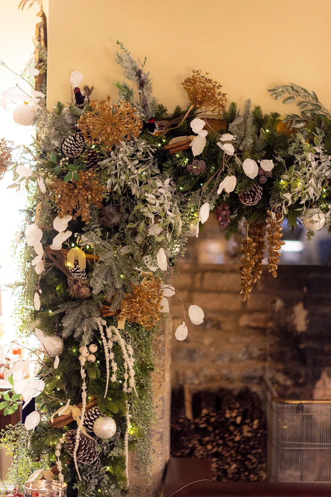 Decorative Christmas garland with gold and white ornaments, pinecones, faux snow-dusted greenery, and string lights hanging above a stone fireplace.
