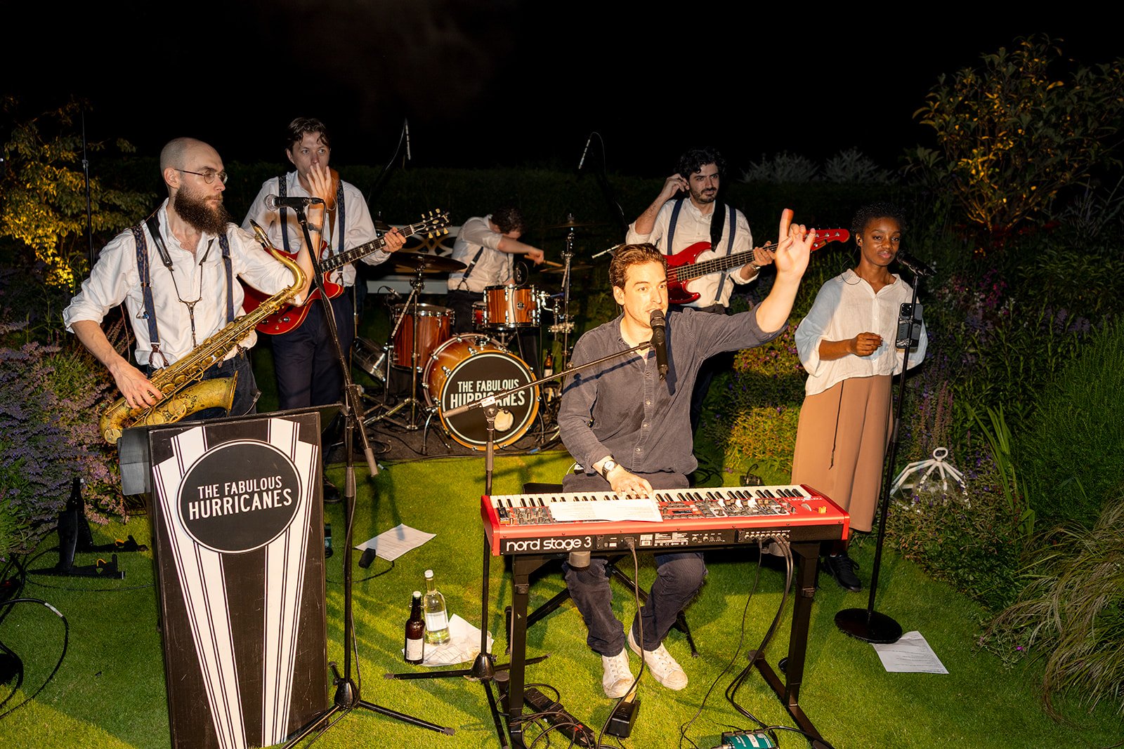 A live band performing outdoors at night surrounded by greenery, with a banner displaying "The Fabulous Hurricanes." The band members are playing various instruments including a saxophone, guitar, drums, keyboard, and vocals, with one member singing 