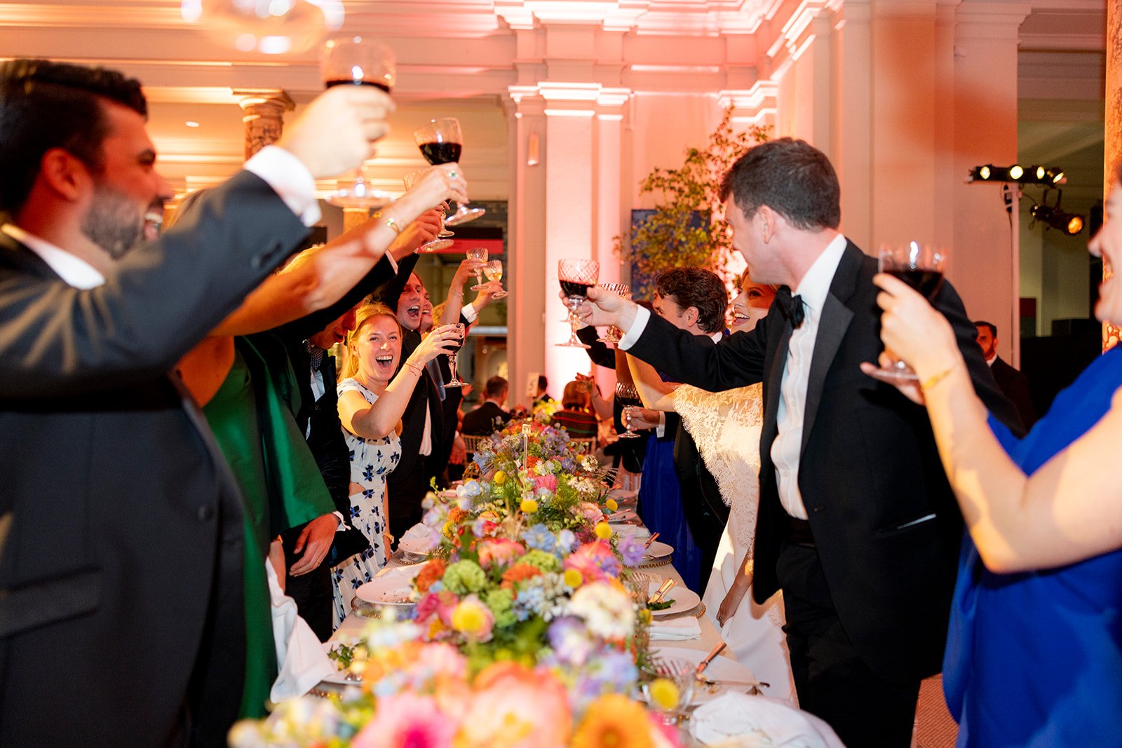 People in formal attire raising glasses in a toast at a wedding reception with floral centerpieces on the table.
