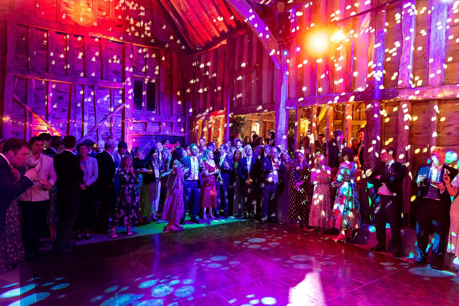 A large group of people dressed in formal attire at an indoor event with colorful stage lighting and wooden walls.