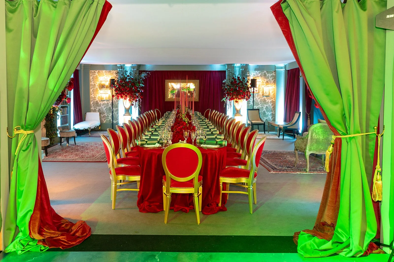 Elegant banquet room decorated with red and gold accents, featuring a long dining table set with glassware, candles, and red floral centerpieces. Green velvet curtains frame the entrance, and the room includes vintage furniture and floral arrangement