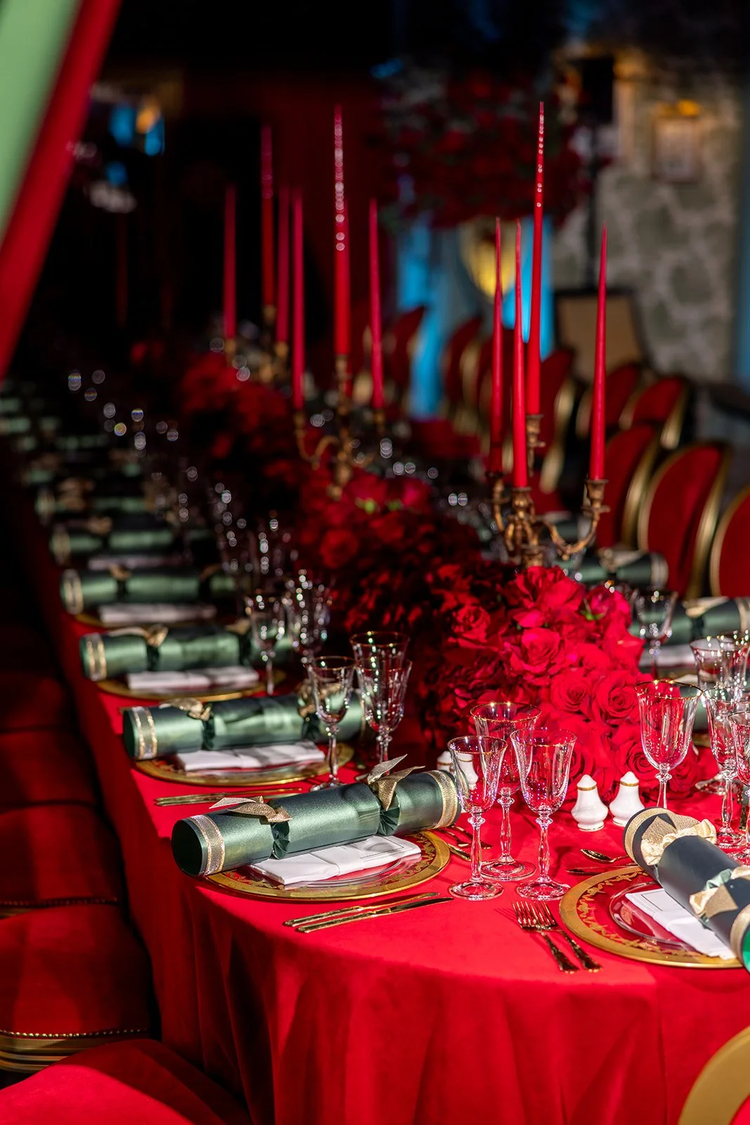 A long banquet table decorated for a celebration with red candles, red roses, green napkins, white plates, gold cutlery, and various glasses, set against a rustic stone wall.