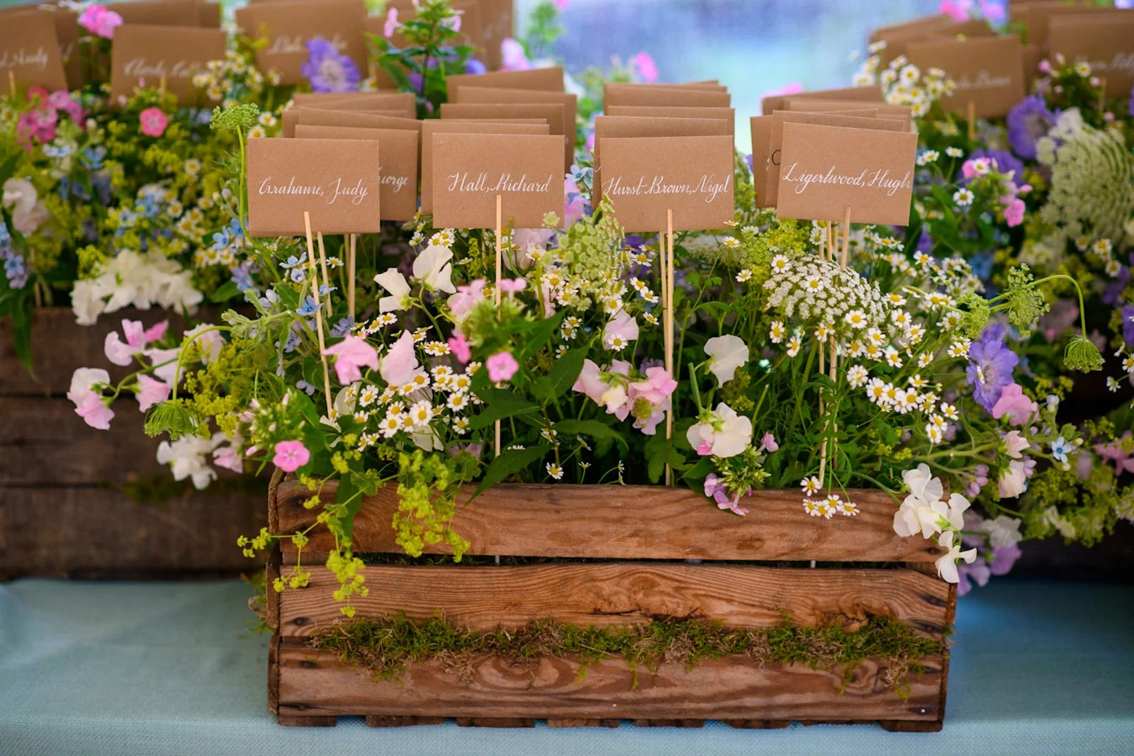 Arrangement of small, colorful flowers with handwritten style table cards on wooden sticks, placed in a wooden planter box.