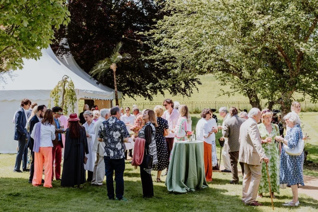 People gathered outdoors near a white event tent, socializing and enjoying drinks under trees on a sunny day.