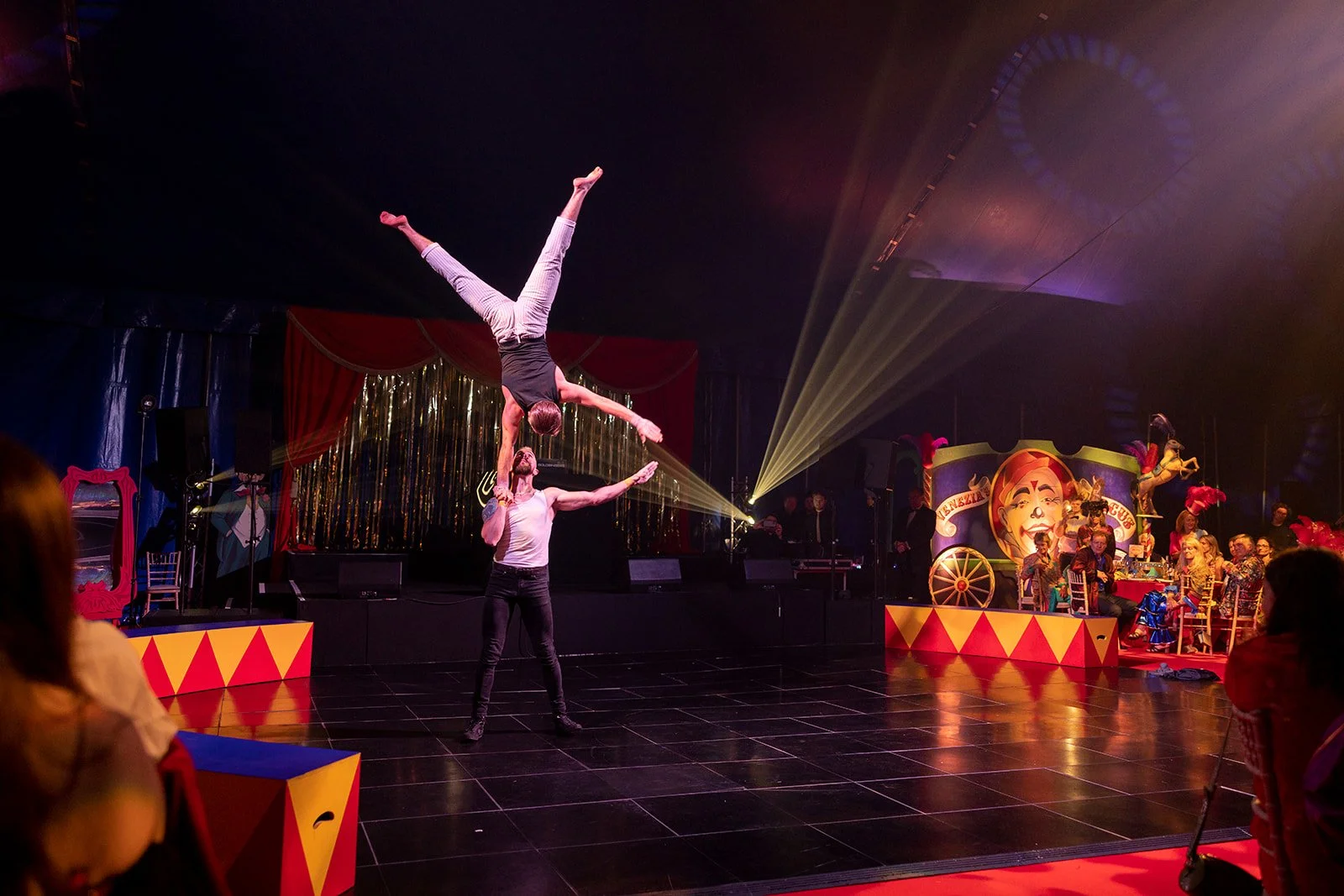 Two acrobats performing a balancing act on a stage decorated for a circus, with one standing and holding the other upside down in a handstand, and circus-themed decorations in the background.