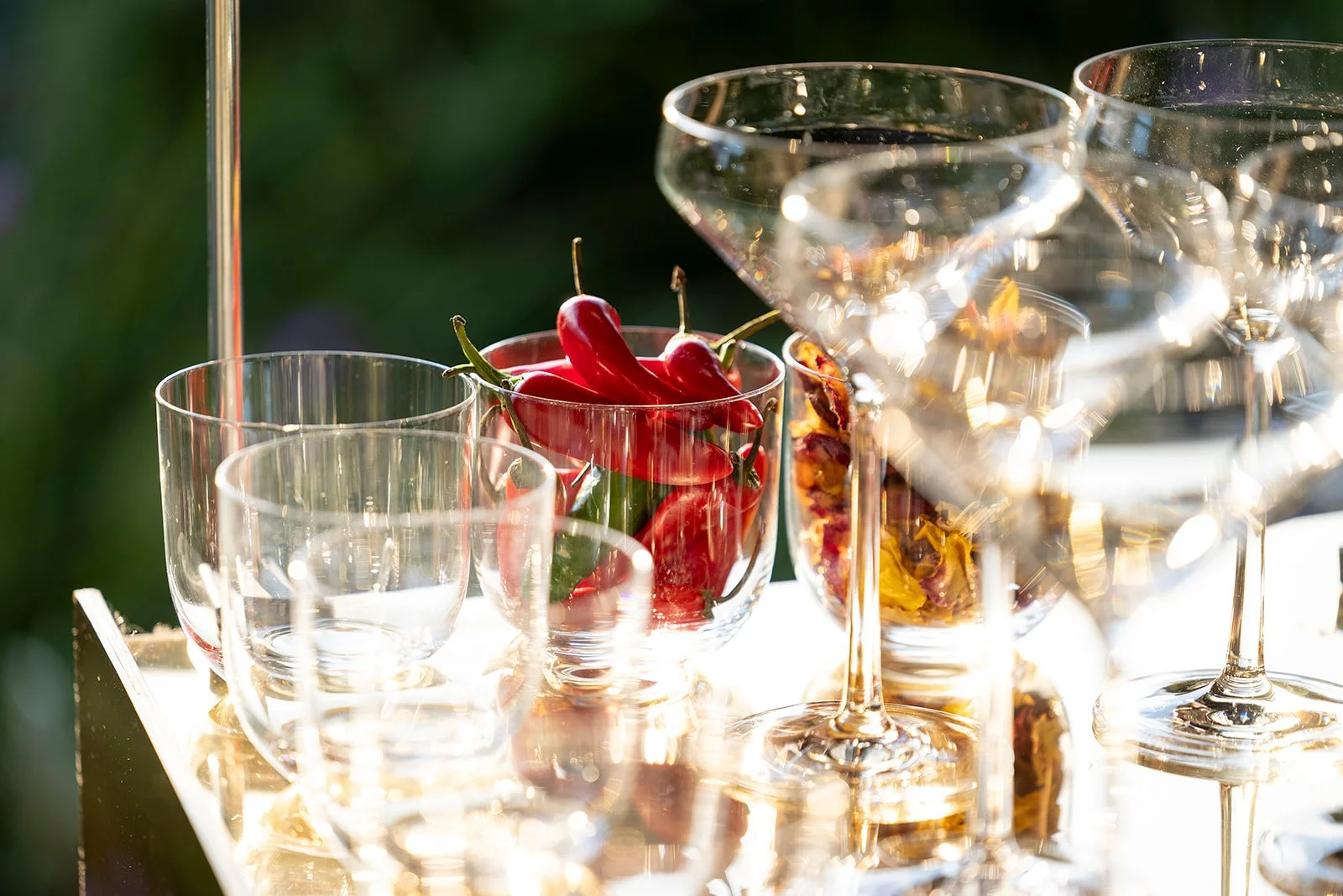 Various empty and filled wine glasses, with one filled with red chili peppers, on a bright outdoor table.