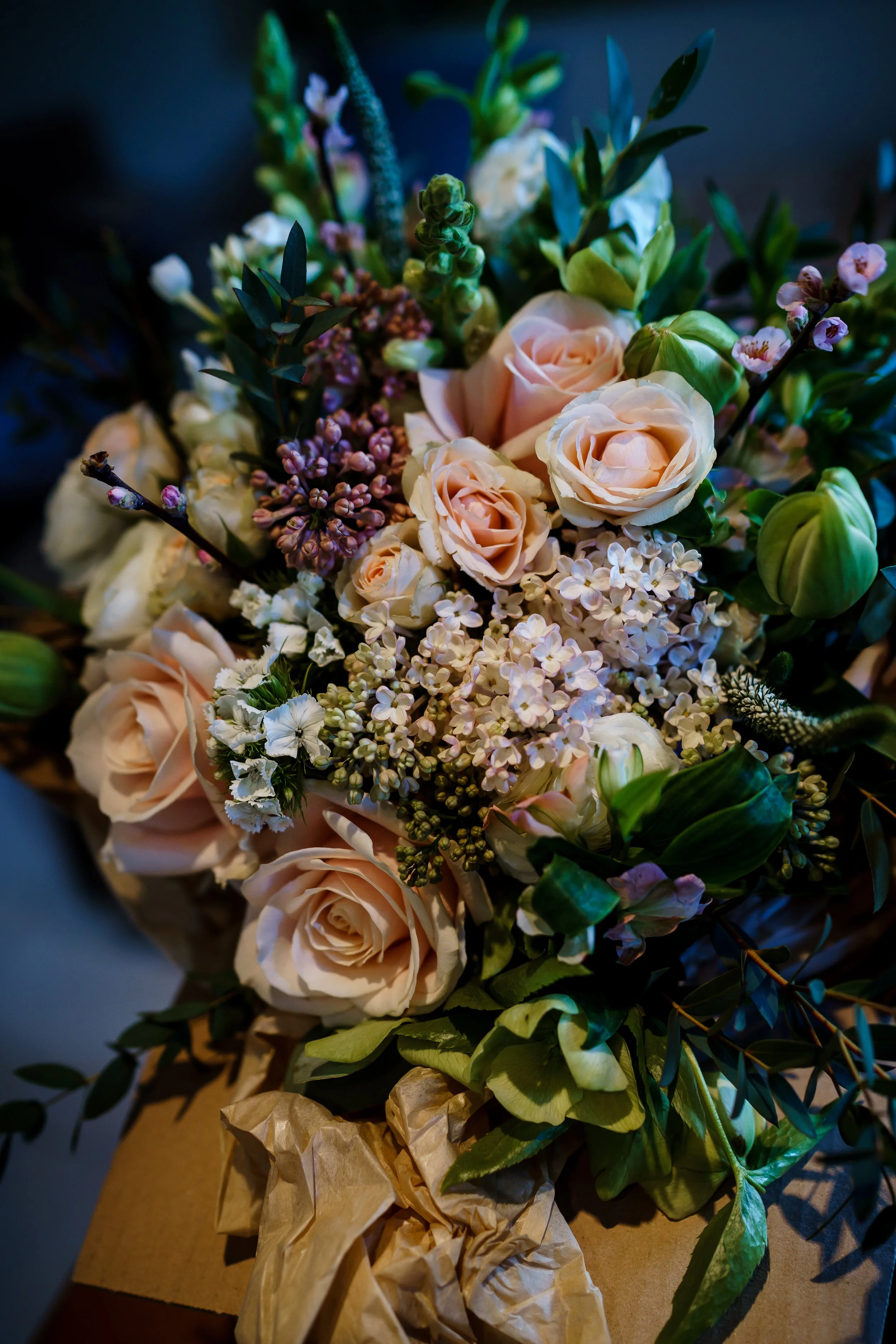 A colorful bouquet of various flowers including pale pink roses, small pink and white flowers, and green foliage, wrapped in brown paper.