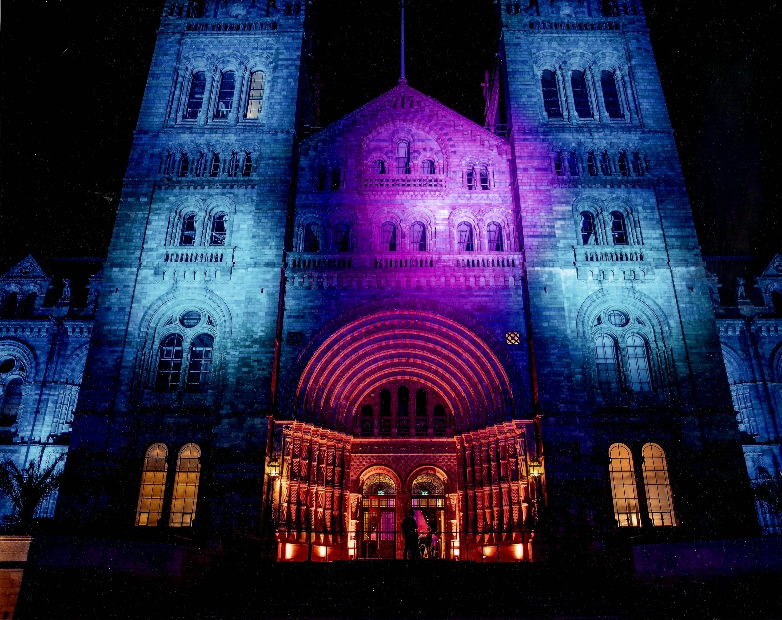 The facade of a large cathedral illuminated with colorful lights, predominantly blue and purple, at night. The building features Gothic architecture with tall arched windows and elaborate details.