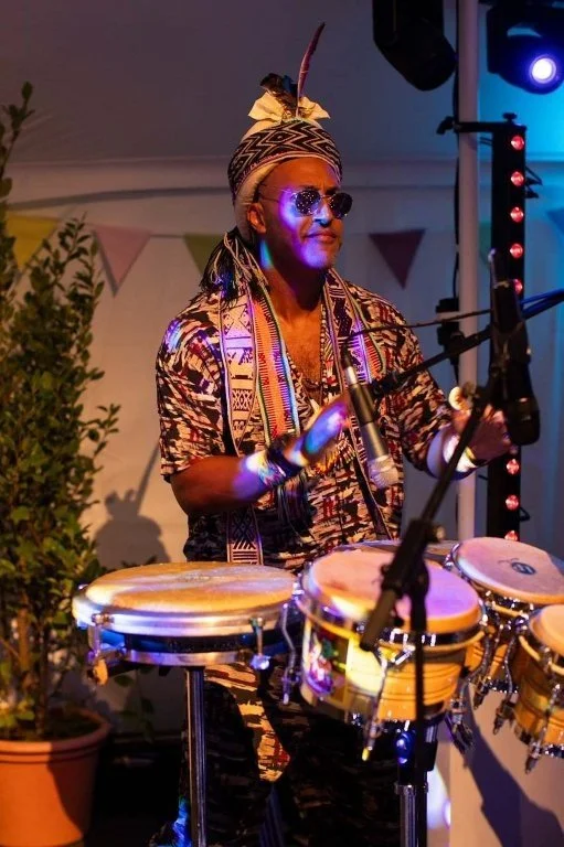 A man playing percussion instruments at a lively event. He is wearing sunglasses, a patterned shirt, a headband with a feather, and various accessories, illuminated by colorful stage lights.