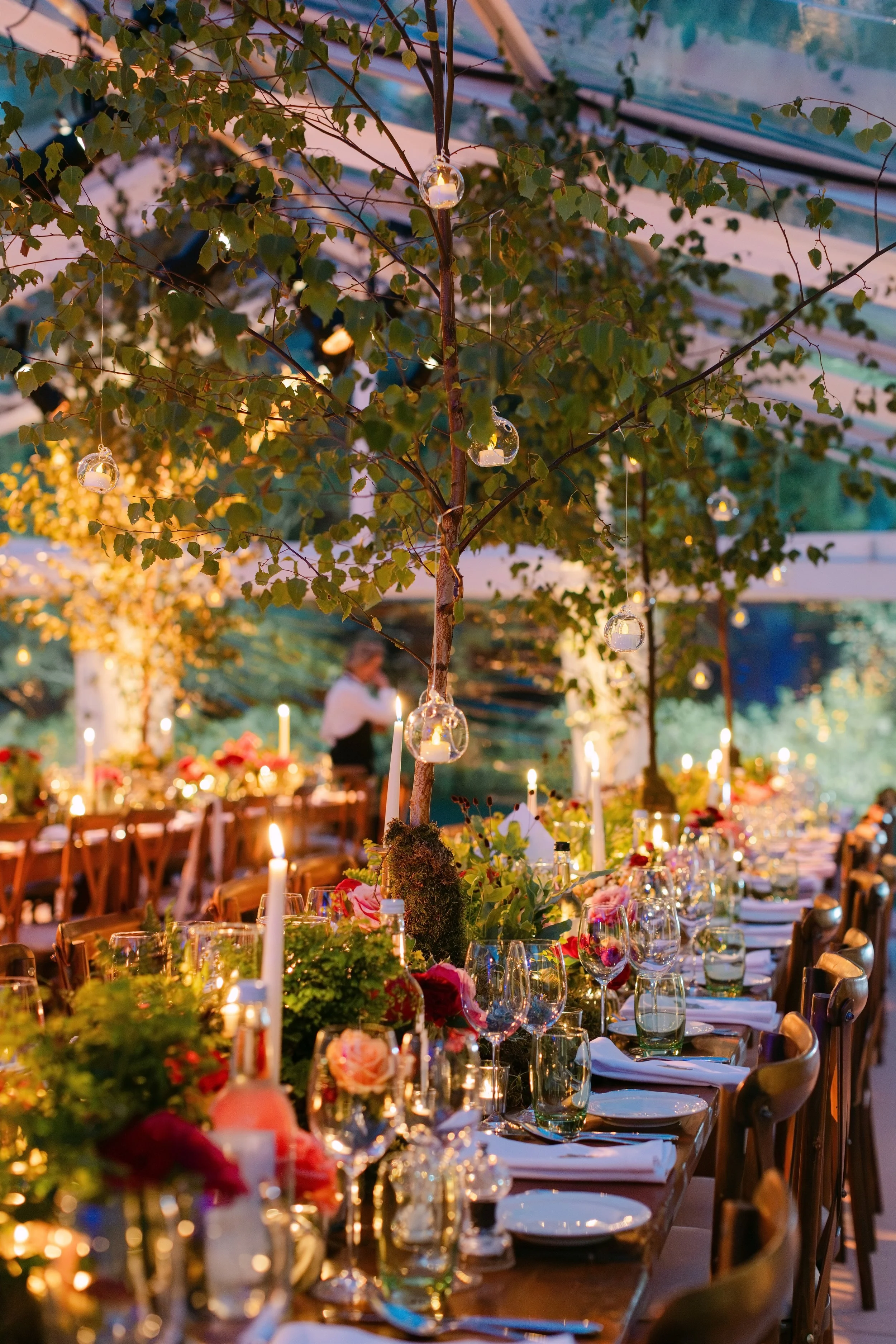 A beautifully decorated outdoor dinner setup under a tree with string lights and hanging glass candle holders, featuring a long table with floral arrangements and dinnerware.