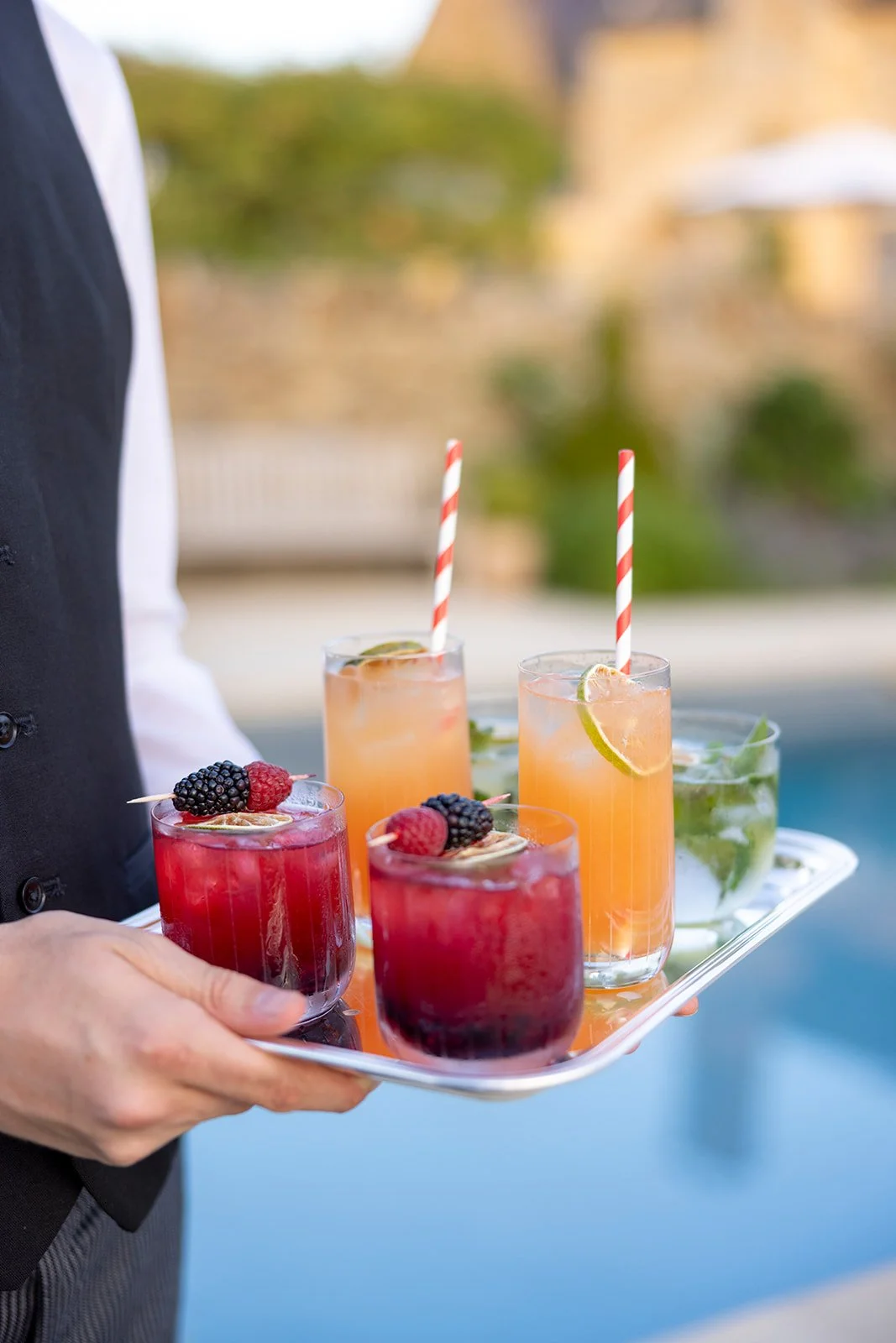 A person in formal attire carrying a tray with colorful cocktails garnished with fruit, set outdoors near a pool.