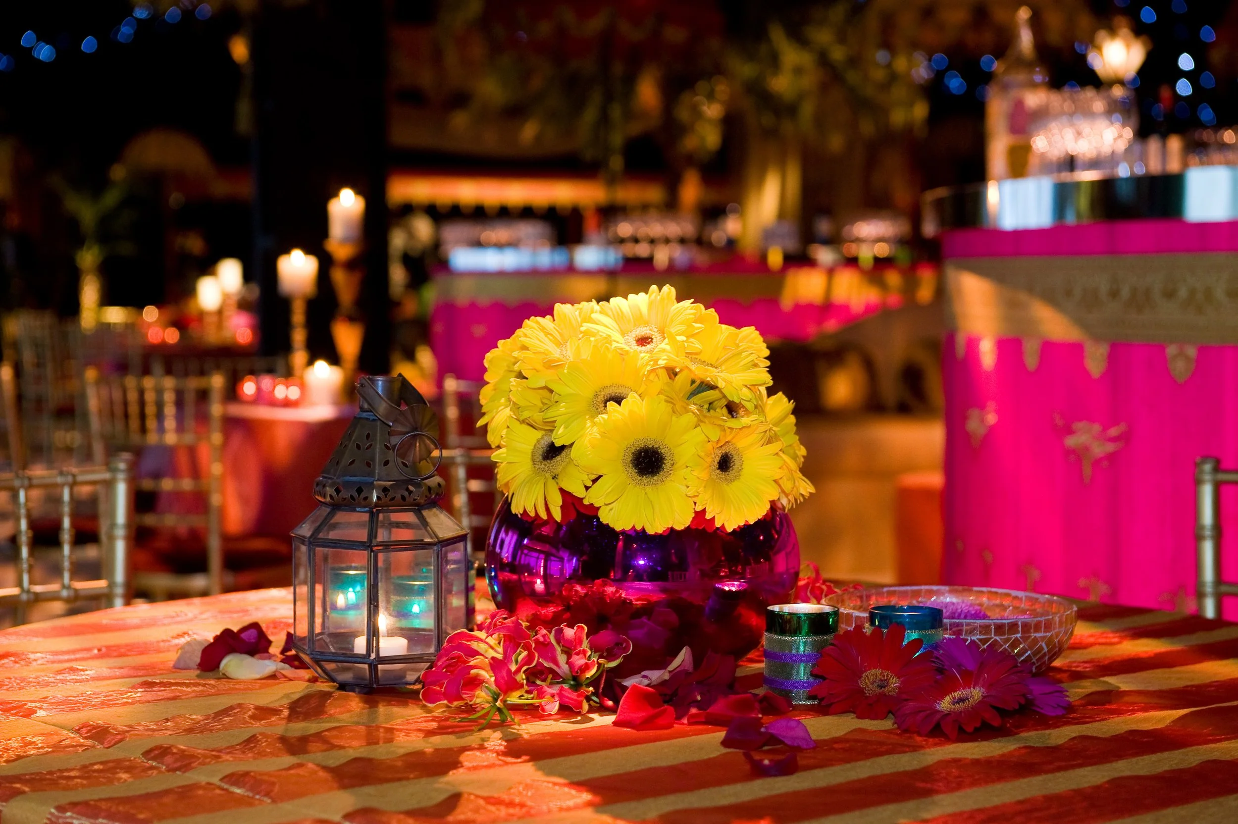 Decorative table setting with a bouquet of yellow flowers in a purple vase, surrounded by colorful candles, scattered flower petals, and small metallic candle holders, in a warmly lit venue with pink and gold accents.