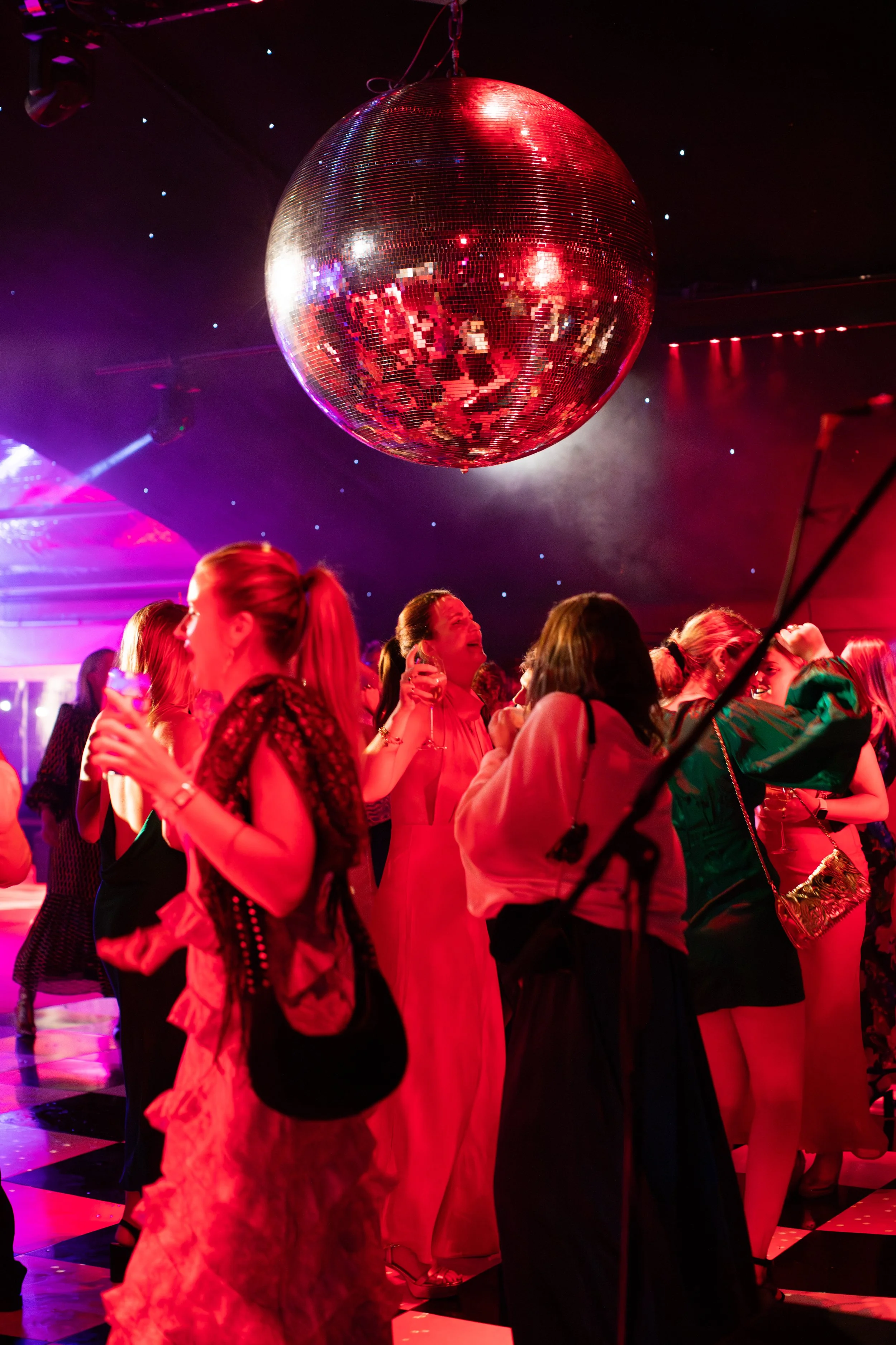 A group of people dancing and socializing at a party or nightclub with a large disco ball overhead, colorful lighting, and a checkered dance floor.
