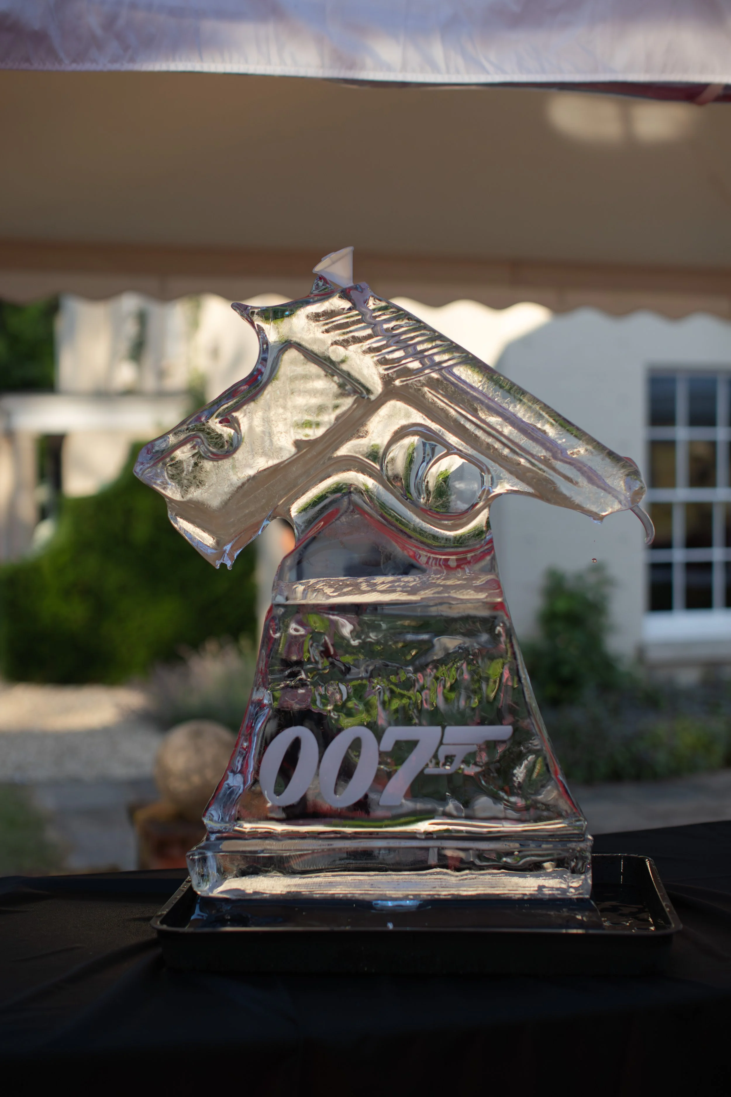 A glass sculpture resembling a trophy with the number "007" etched on it, sitting on a black tray outdoors with a white house and greenery in the background.