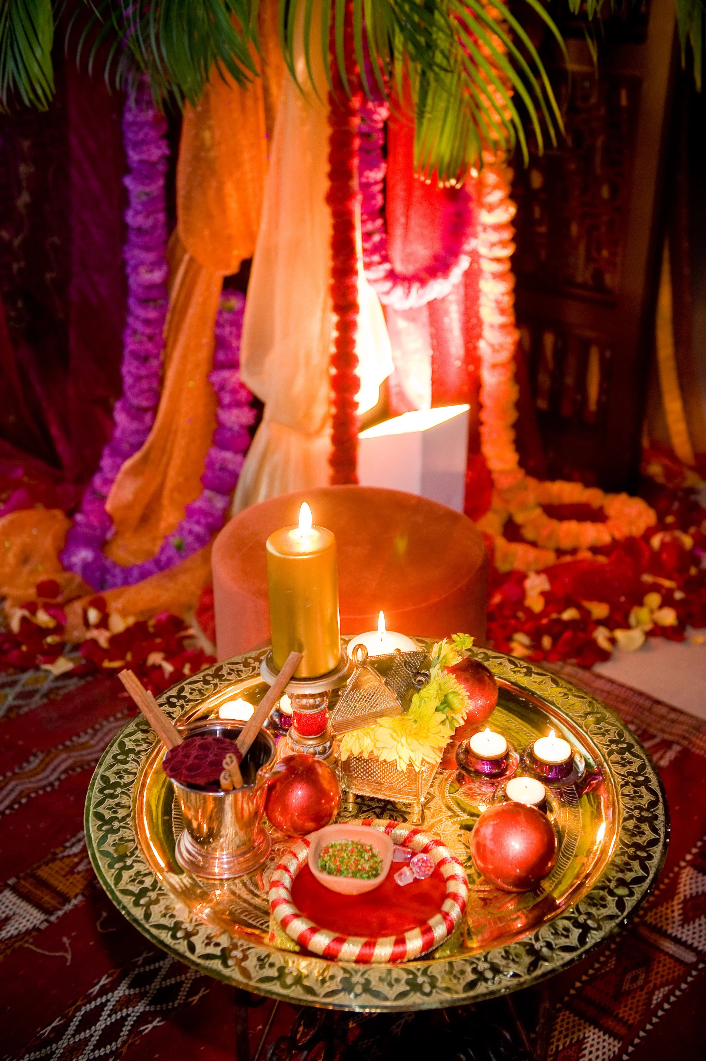 Hindu ceremonial setup with lit candles, pomegranate fruits, decorative items, and flower garlands in the background, for a religious or cultural celebration.