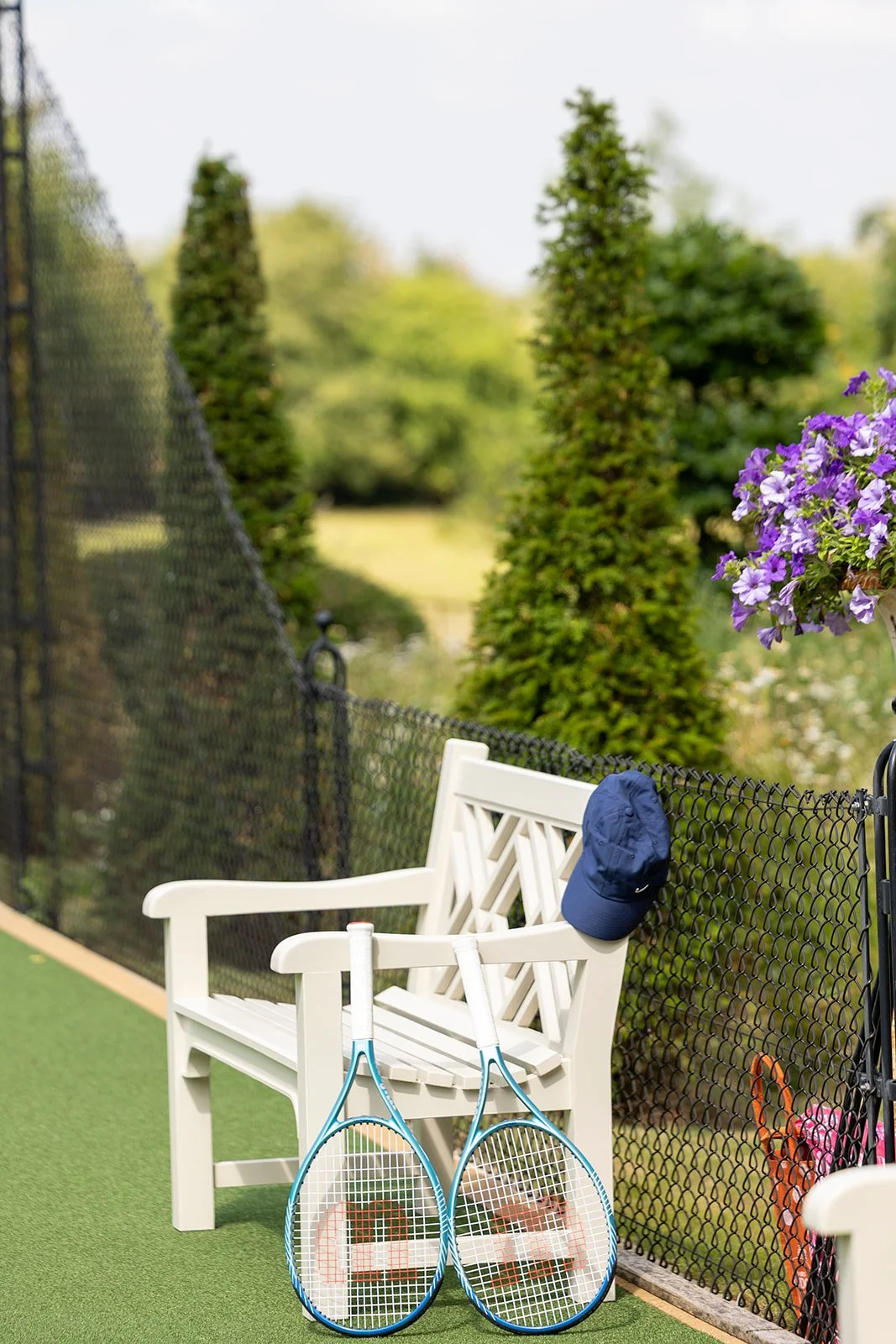 A white bench with a blue cap hanging on the backrest, two tennis rackets leaning against it, on a tennis court with green artificial grass. Tall evergreen trees and purple flowers are visible in the background.