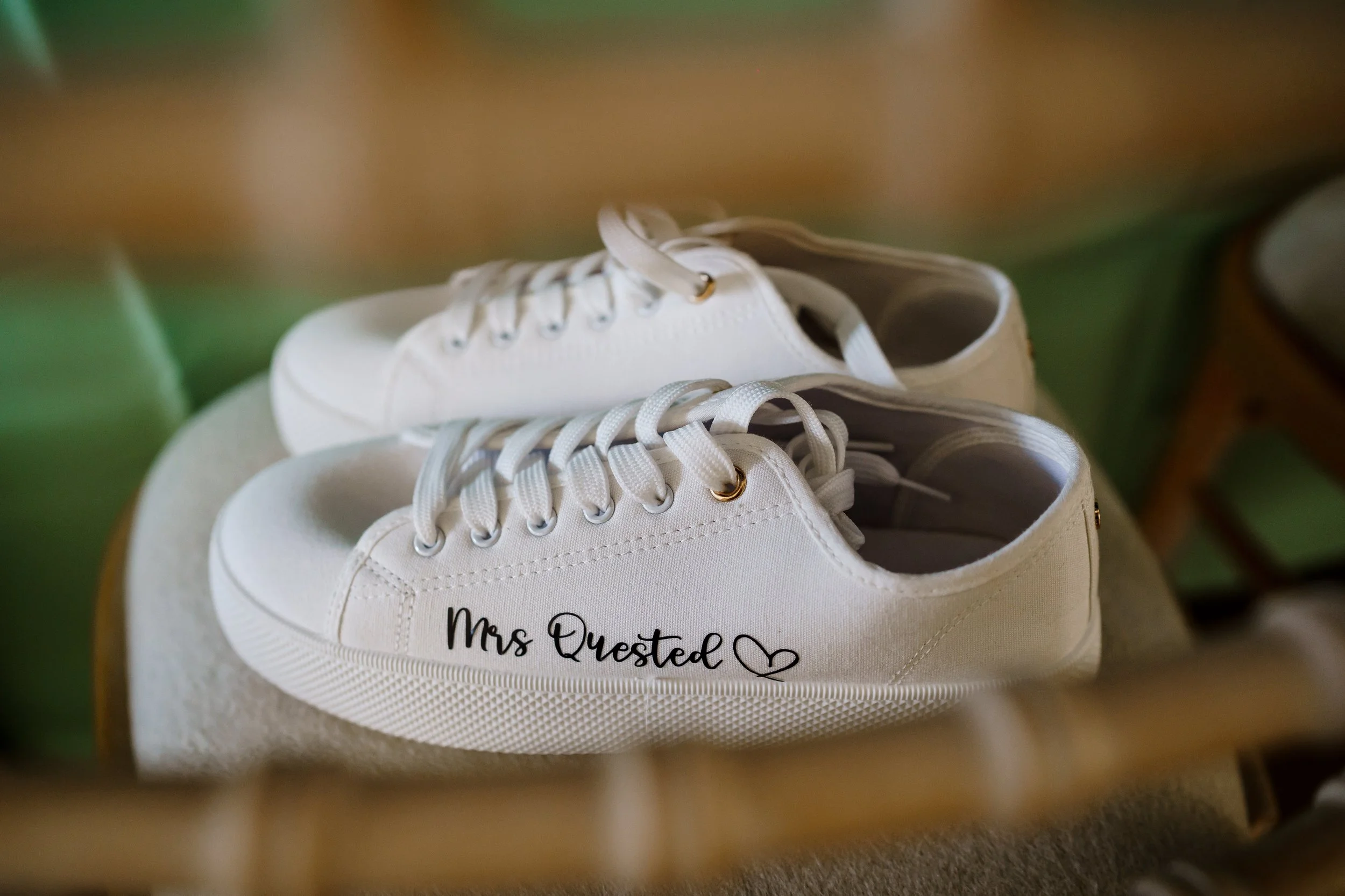 Close-up of white sneakers with custom text reading 'Mrs. Quested' and a heart on the side, resting on a surface with a blurry background.