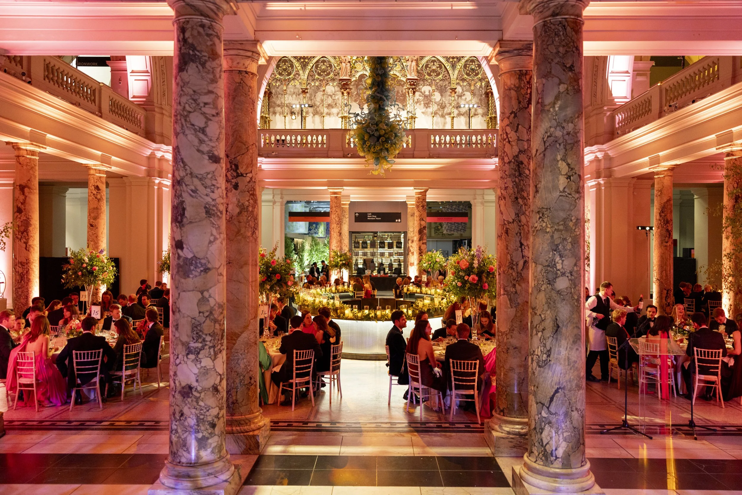 Elegant ballroom decorated for a formal event with marble columns and pink lighting, filled with people seated at round tables with floral centerpieces and a bar area in the background.