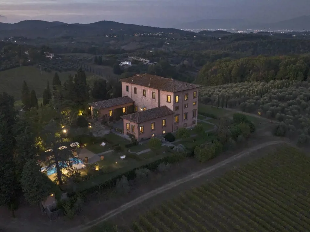 A large, multi-story house in a rural area with illuminated windows, surrounded by trees, vineyards, and a garden with a swimming pool, in a valley during evening or dusk.