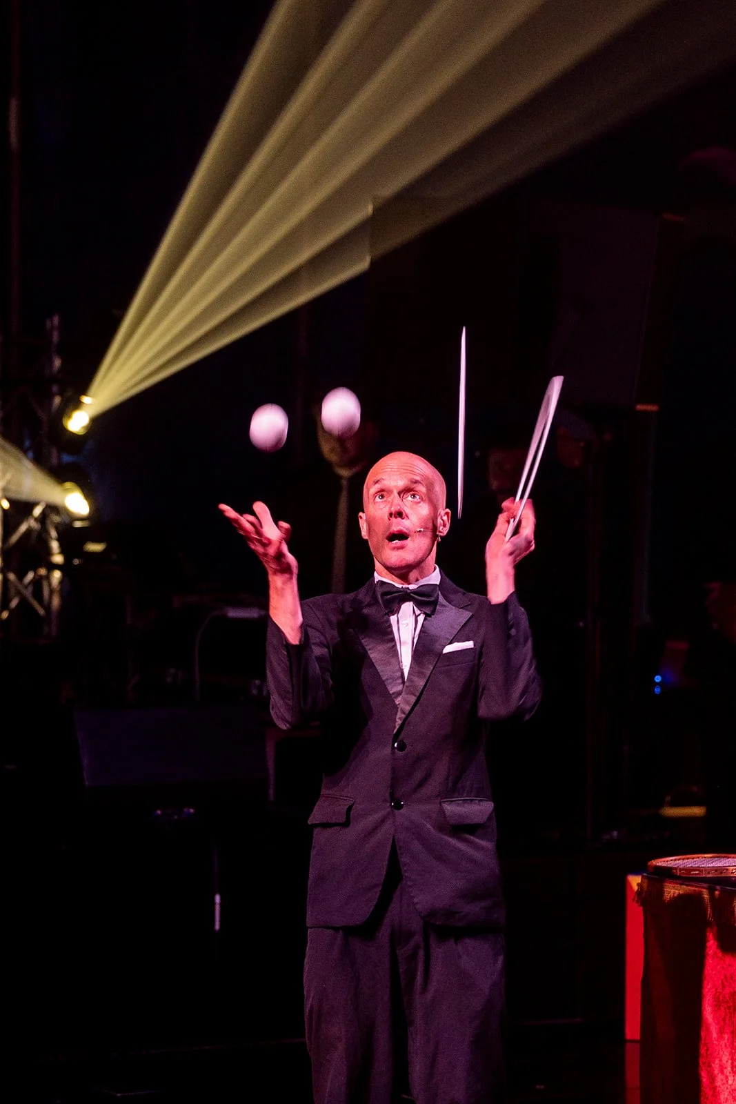 A mime artist dressed in a tuxedo performs with a baton on stage, with yellow stage lights and hanging spherical lights in the background.