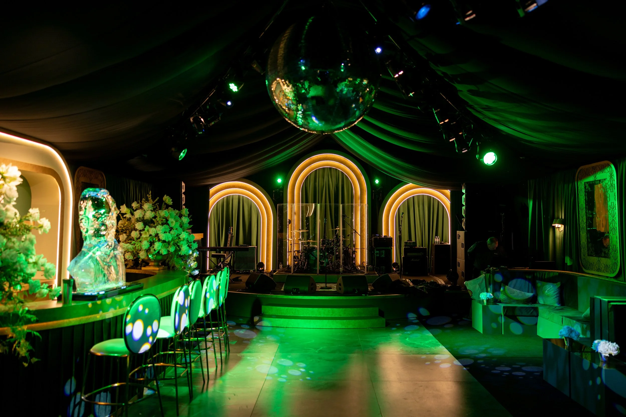 A dimly lit event stage decorated with green and gold lighting, featuring a large disco ball hanging from the ceiling, musical instruments on stage, and a dance floor in front.
