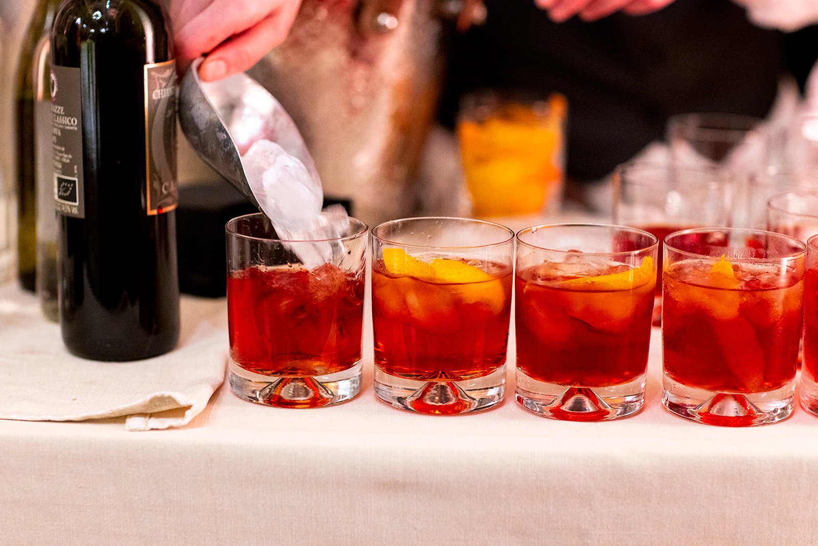 Four glasses filled with red-colored cocktail with ice and orange slices, a person pouring a drink from a bottle, and a black bottle on a table with a white tablecloth.