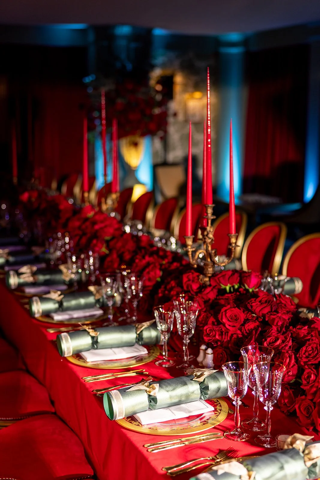 A lavishly decorated banquet table with red roses, gold accents, tall red candles, glassware, and neatly arranged cutlery in an elegant setting, possibly for a celebration or special event.