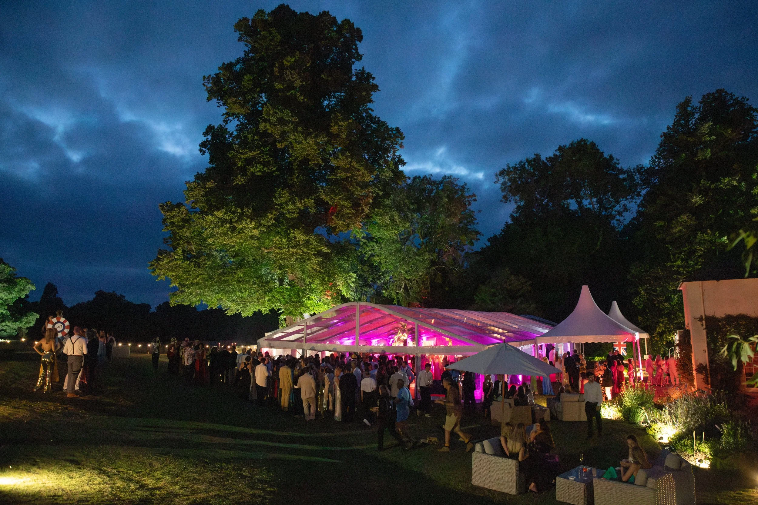 People gathered outdoors during evening at an event with illuminated white tents and string lights, under a dark cloudy sky surrounded by trees.