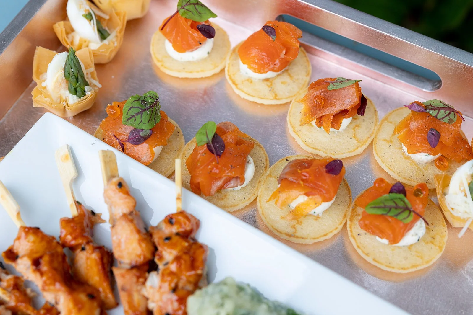 Assorted small appetizers including smoked salmon on rounds with cream and microgreens, and skewers of glazed chicken.