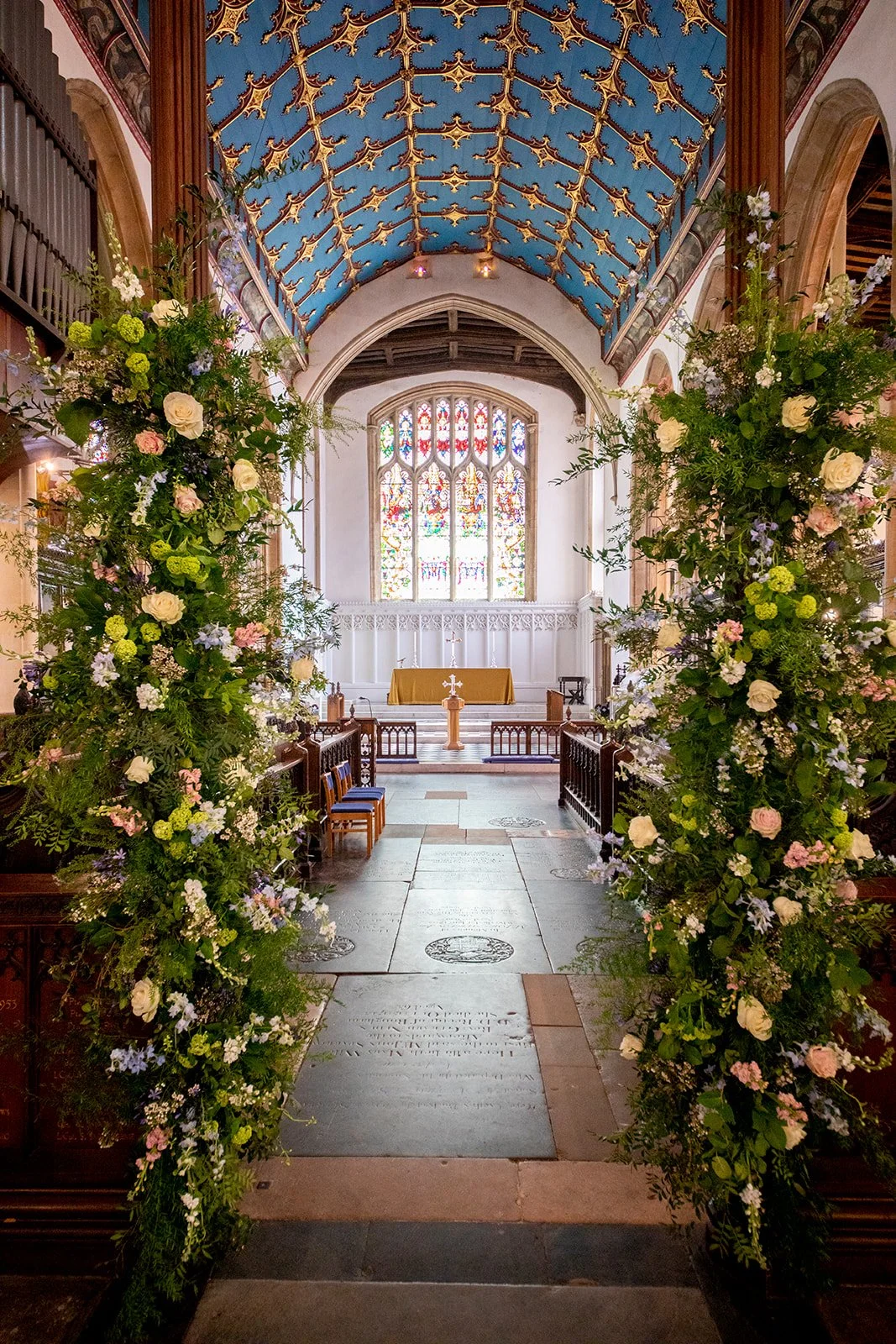 Interior of a church decorated for a wedding with floral arrangements on either side of the aisle, stained glass window behind the altar, and a blue, gold-decorated vaulted ceiling.