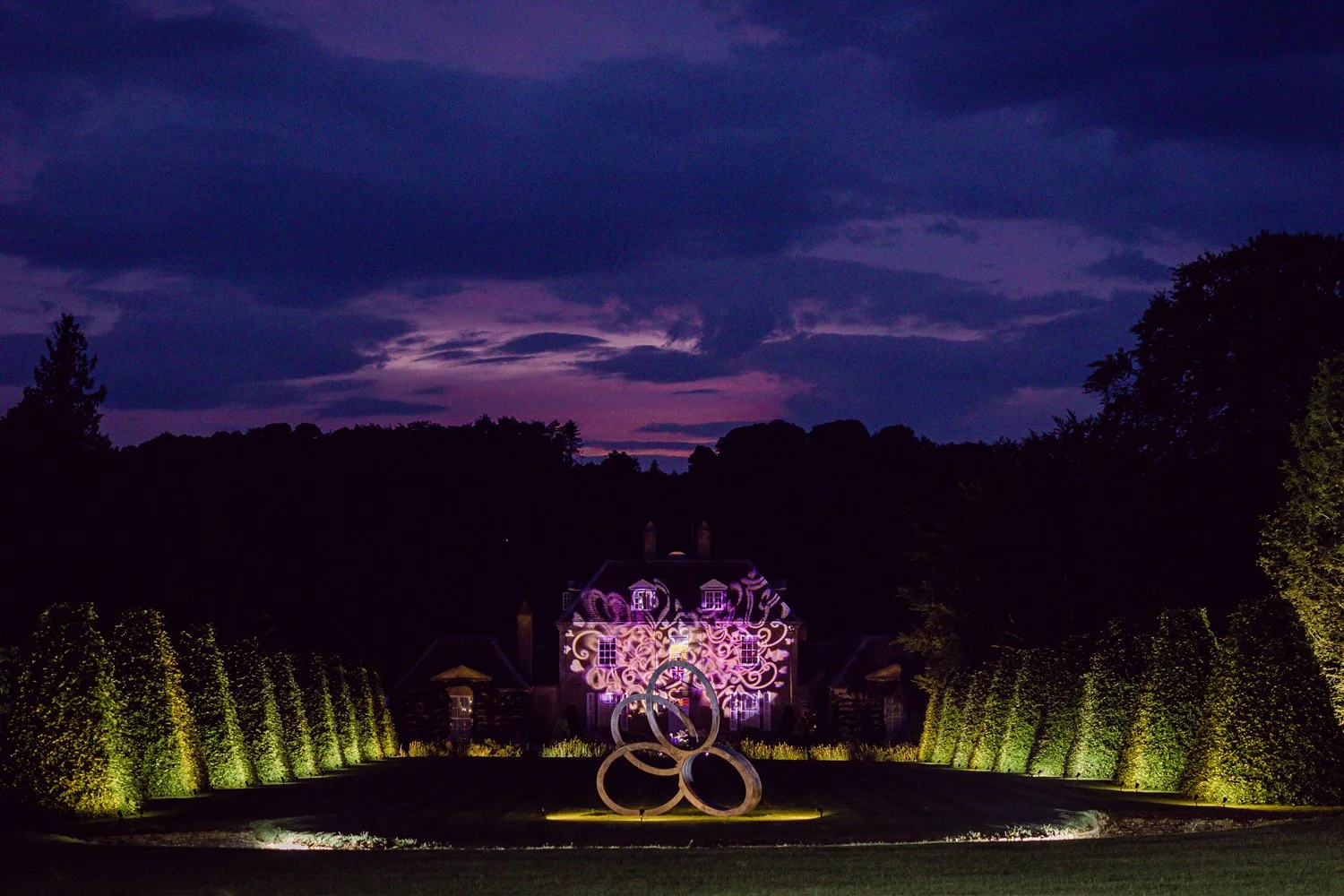 A large illuminated house with decorative lighting projections on its facade, surrounded by green hedges, with a modern sculpture of three intertwined rings in front, taken at dusk or night.