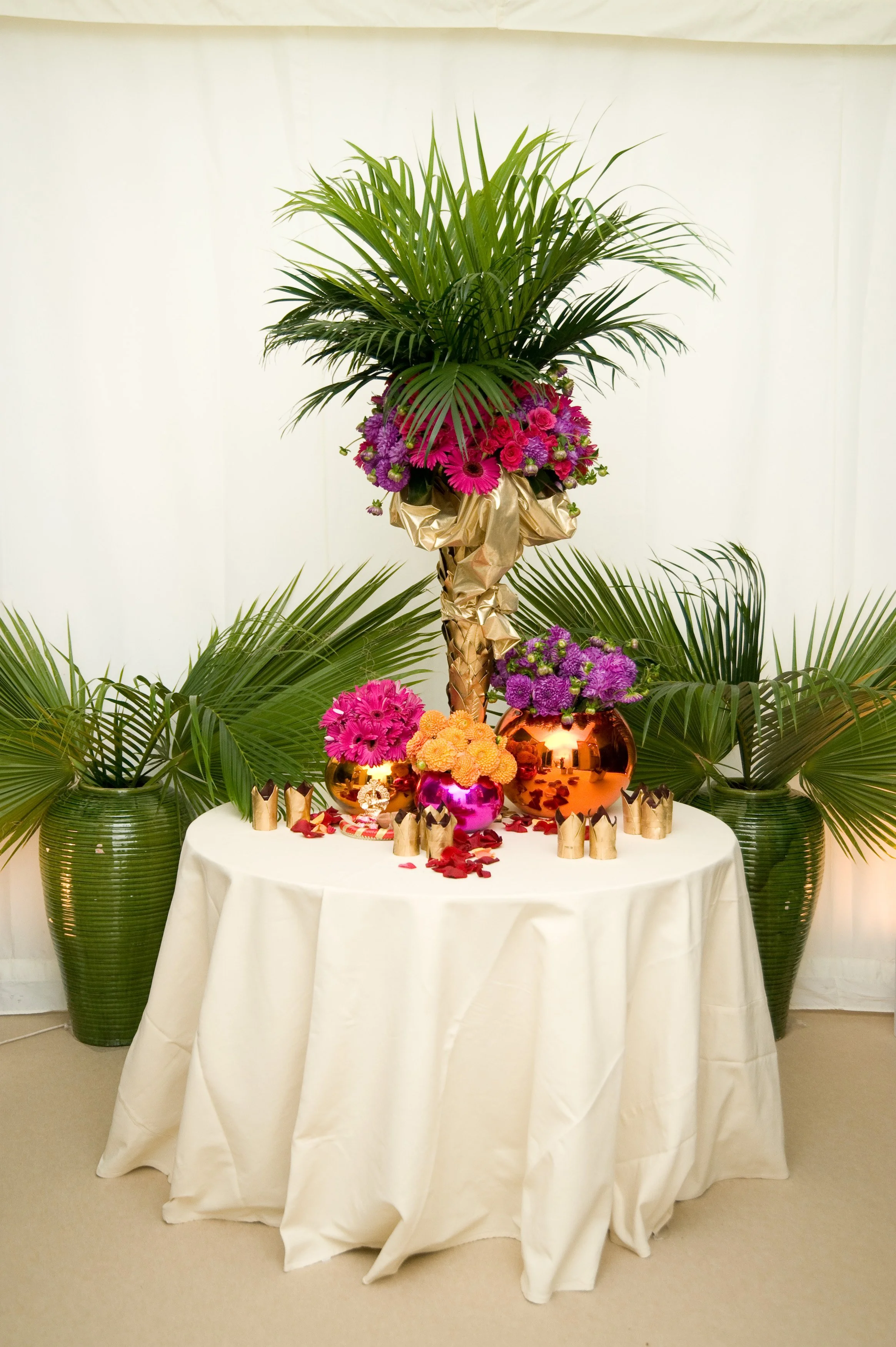 A floral display on a round table with a white tablecloth, featuring pink, purple, and orange flowers, surrounded by gold vases and small crown-shaped decorations, with large green palm leaves in the background.