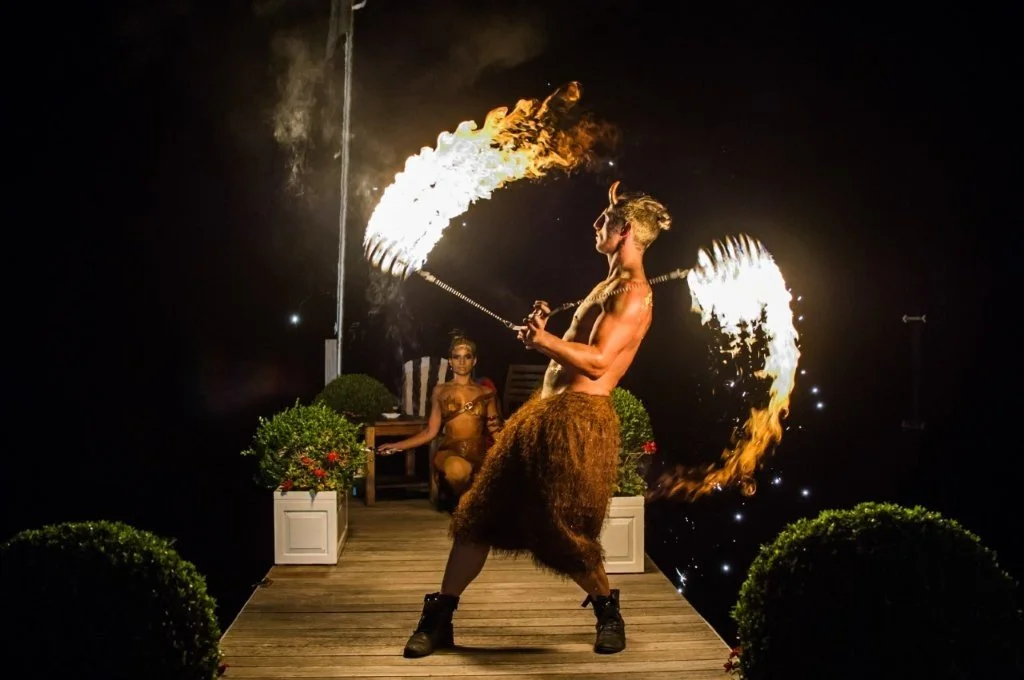 A fire performer at night on a wooden stage, spinning flaming poi to create arcs of fire, with a woman seated in the background.