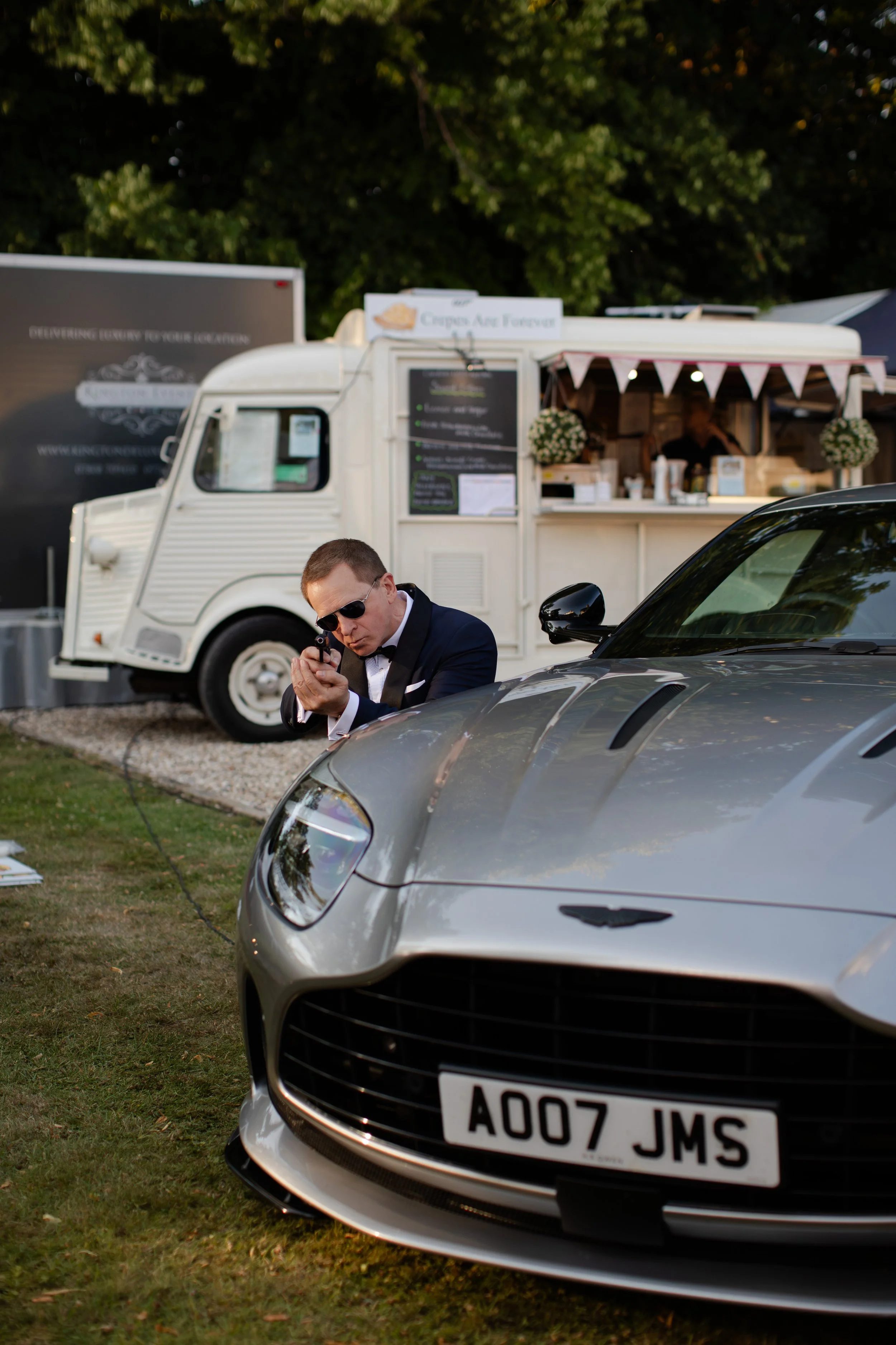 A man in a tuxedo and sunglasses kneeling next to a silver sports car with a license plate A007 JMS, with a white food truck and display stand in the background at an outdoor event.