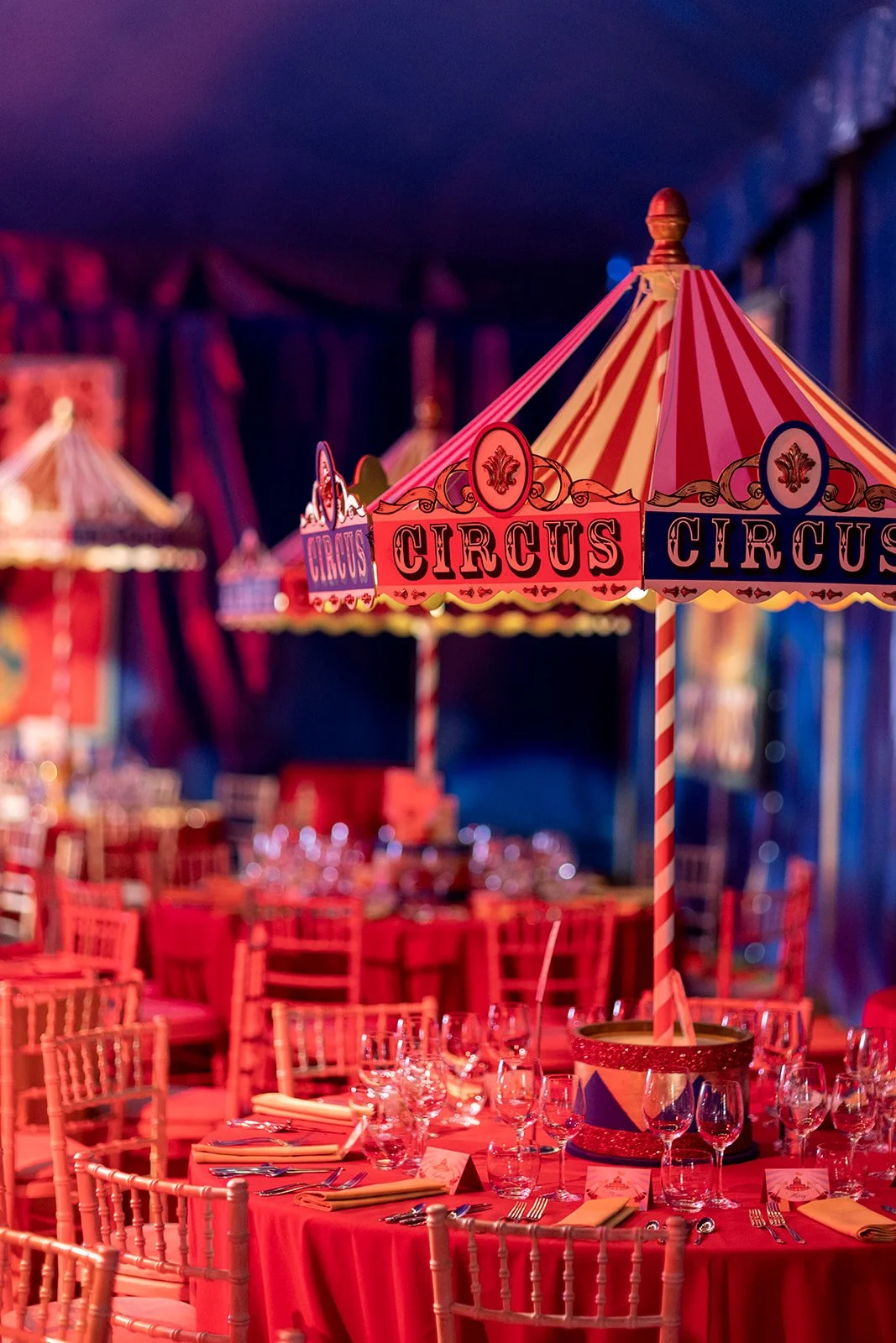A circus-themed event setup with tables decorated in red, with a prominent striped red and yellow circus tent centerpiece, and another similar tent in the background, all inside a dimly-lit venue.