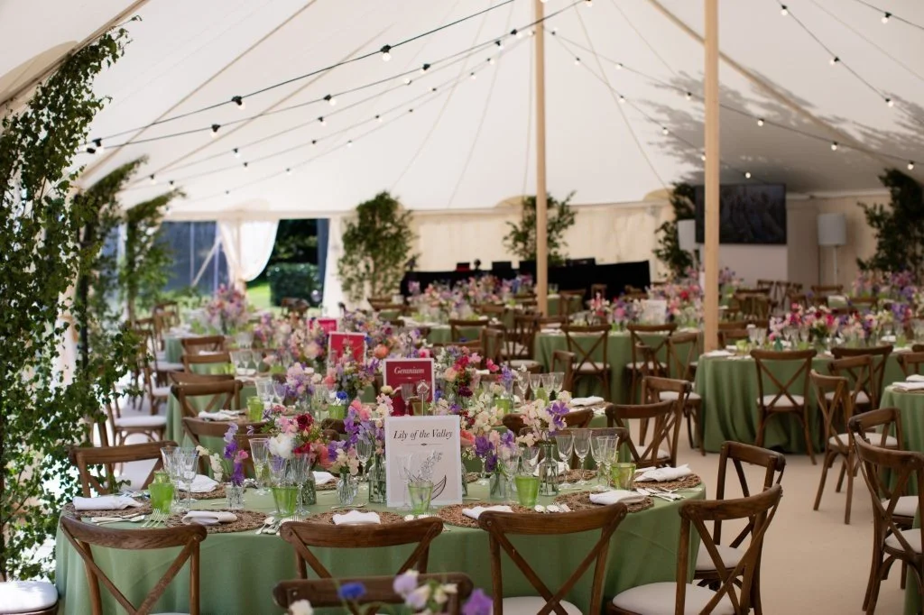 A decorated event tent with multiple round tables covered with green tablecloths, set with glassware, silverware, napkins, and floral centerpieces, illuminated by string lights overhead.